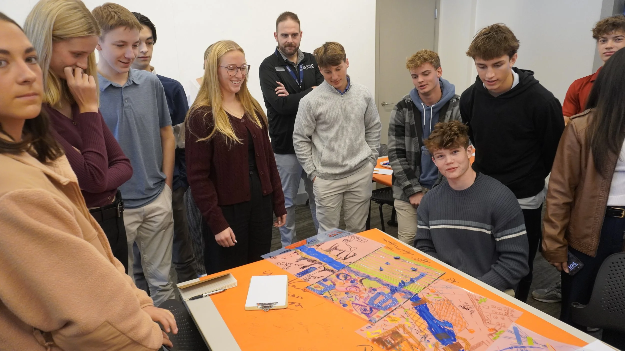 Group of young students and an adult gathered around a table with colorful science project posters, some smiling and looking at the displays.