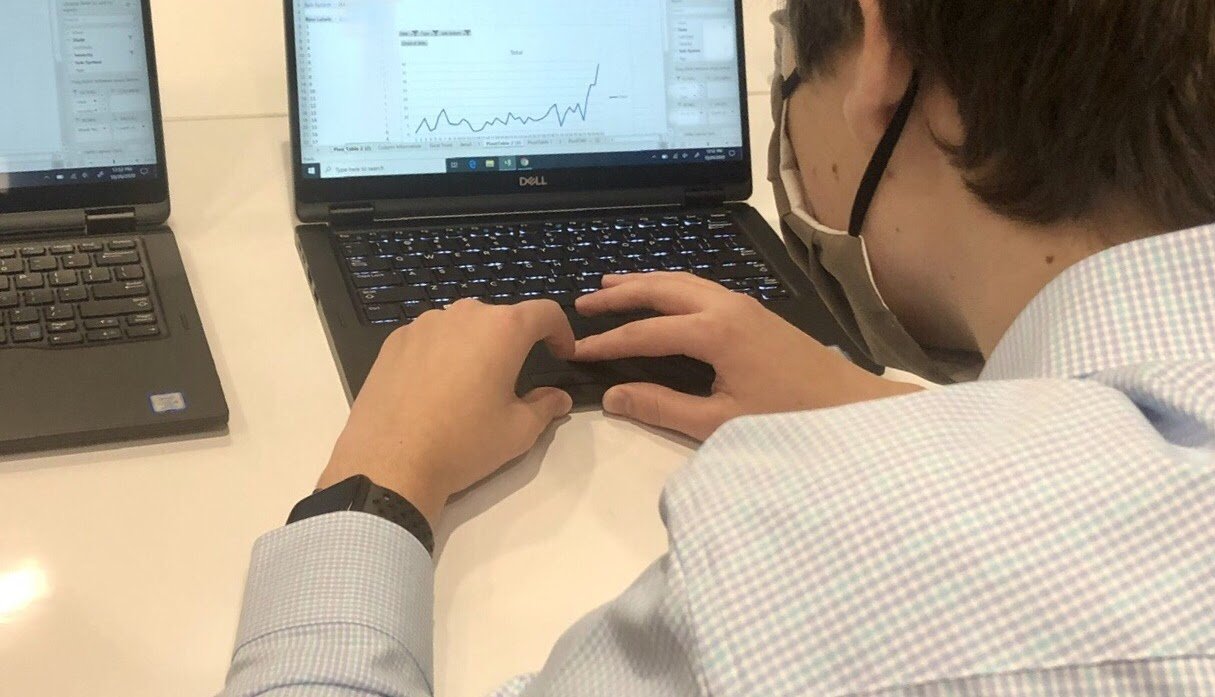 Person wearing glasses and a face mask working on a laptop displaying a graph or chart, with another laptop nearby.