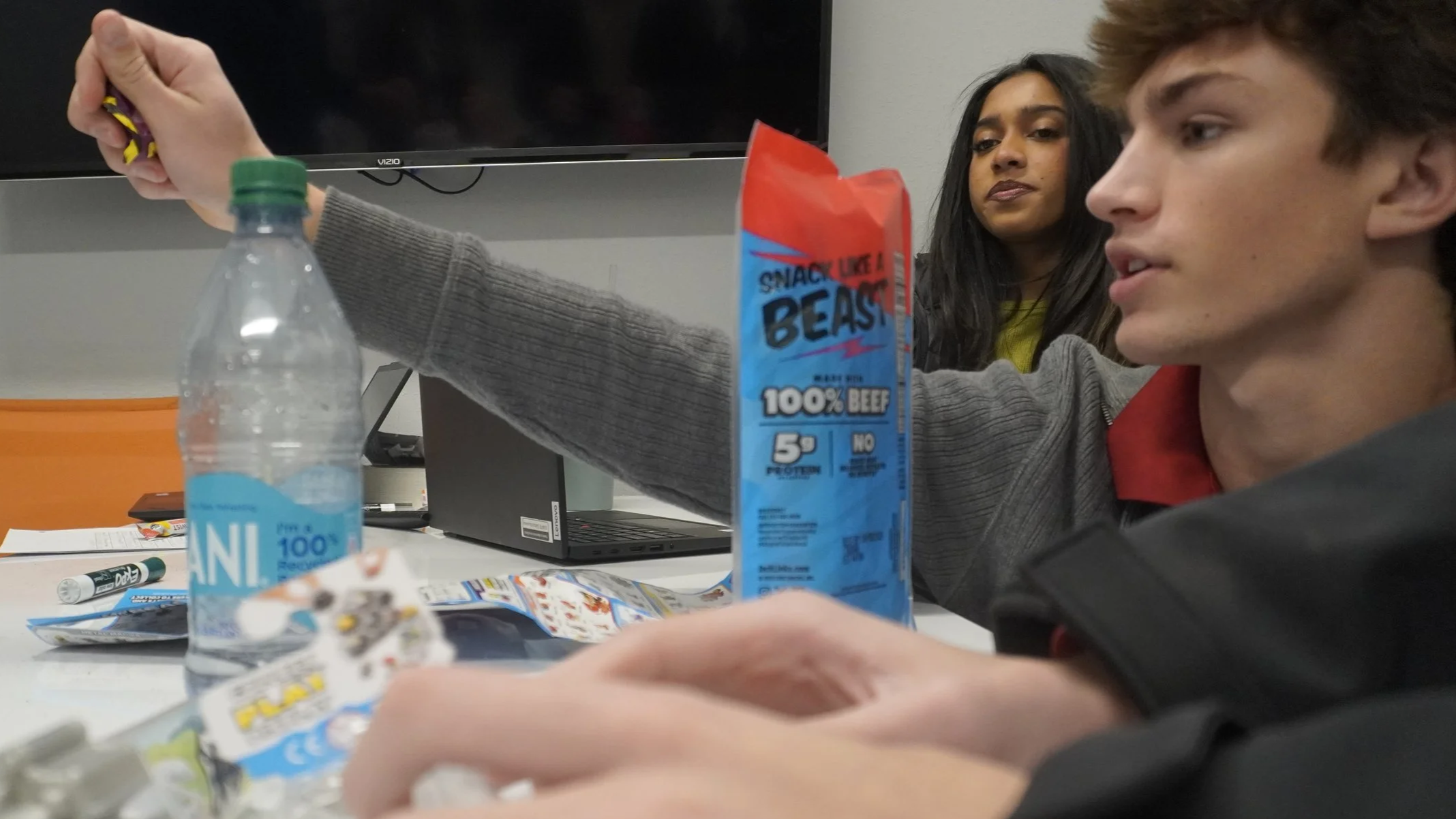 Two young adults, a man and a woman, sit at a table with snacks and a water bottle, in a room with a TV and a computer. The man is reaching for a snack bag, while the woman looks on.
