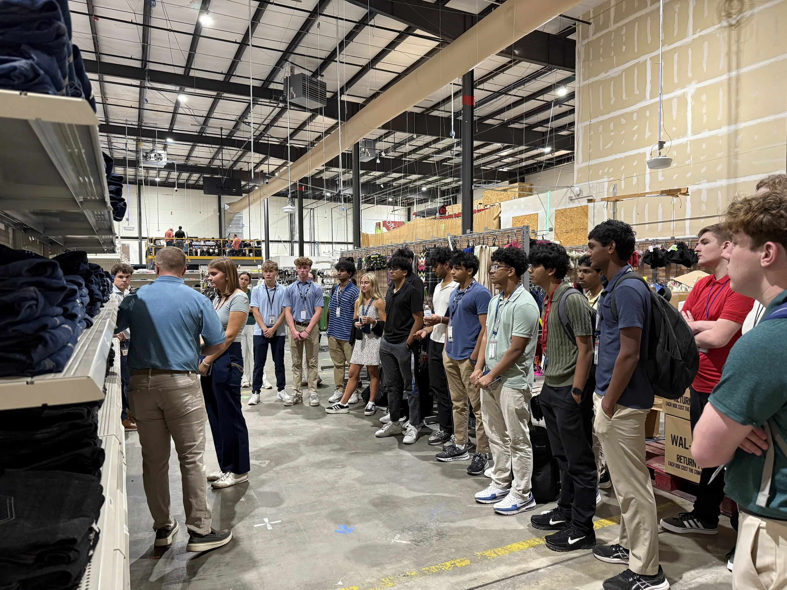 A group of young people, wearing lanyards, listening to a man in a blue shirt speaking inside a retail store that sells clothing.