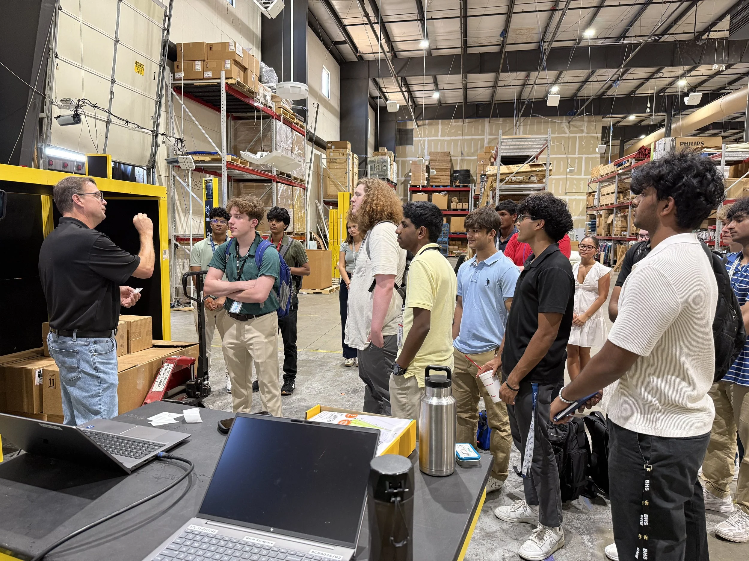A group of young people listening to a man giving a presentation inside a warehouse. The warehouse has shelves loaded with boxes and equipment, and a table with laptops and water bottles in the foreground.