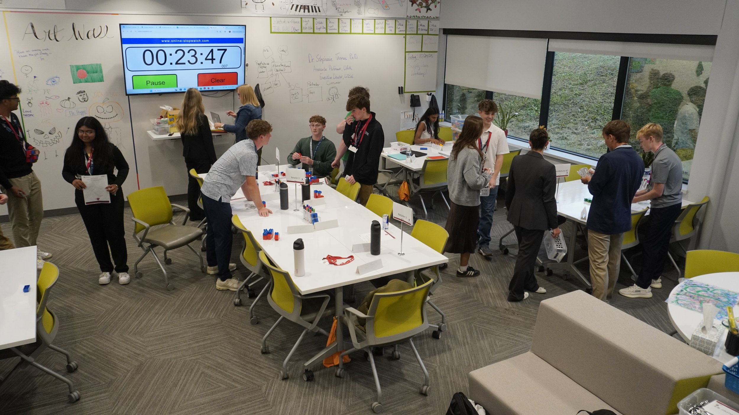 Students participating in a classroom activity involving colorful blocks, with some students standing and engaging at tables, others browsing materials, and a large digital timer on the wall displaying 00:23:47, in a well-lit room with whiteboard wal