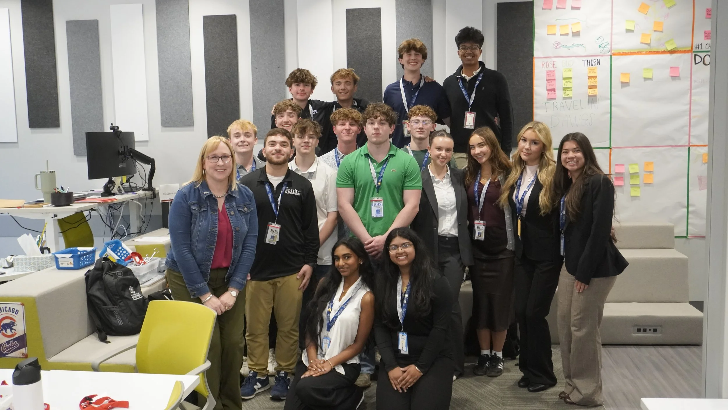 Group of young adults and adults in a classroom or office setting, smiling for a photo, with sticky notes and charts on the wall, and desks with various items in the background.