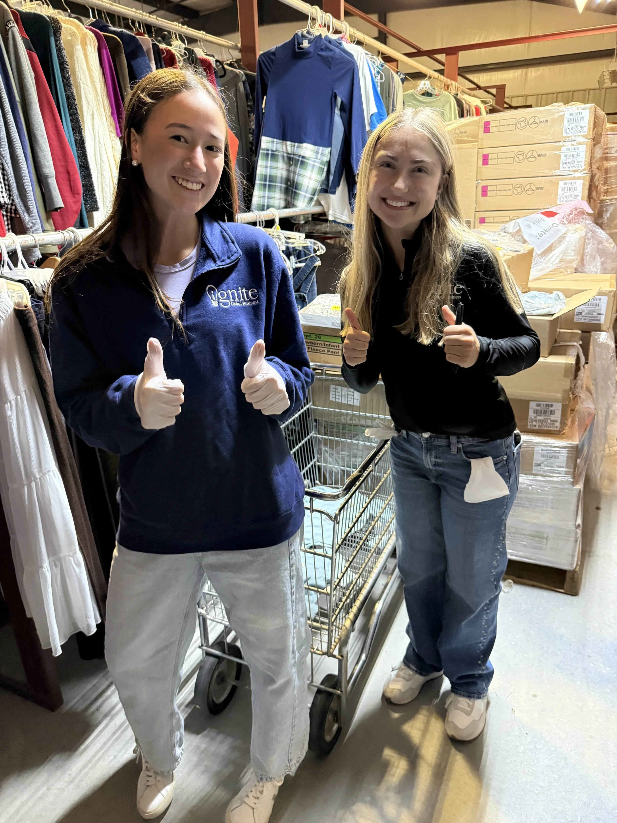 Two women smiling and giving thumbs up inside a warehouse or store with clothing and boxed items around them.