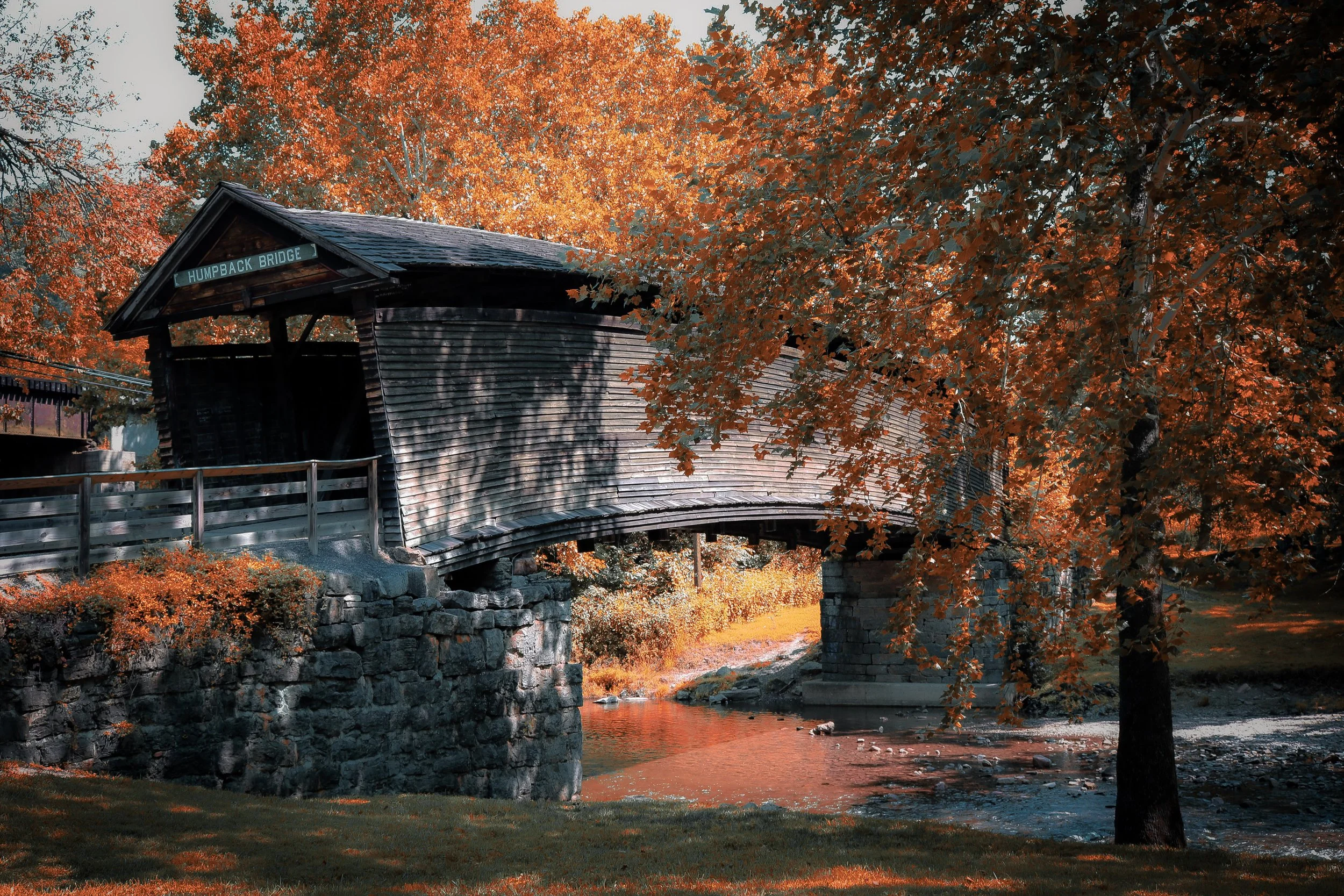 Autumn scene with a covered wooden bridge over a small stream, surrounded by trees with orange and yellow leaves.