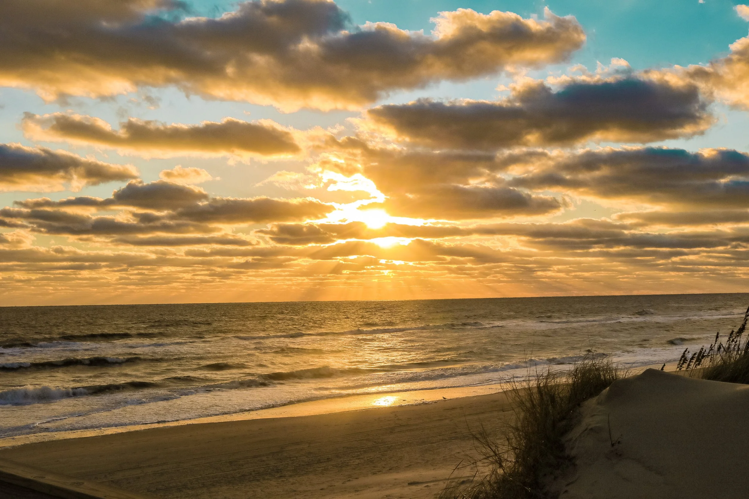Sunset over the ocean with partly cloudy sky, casting golden light on the water and sandy beach below, with some dunes and grass in the foreground.