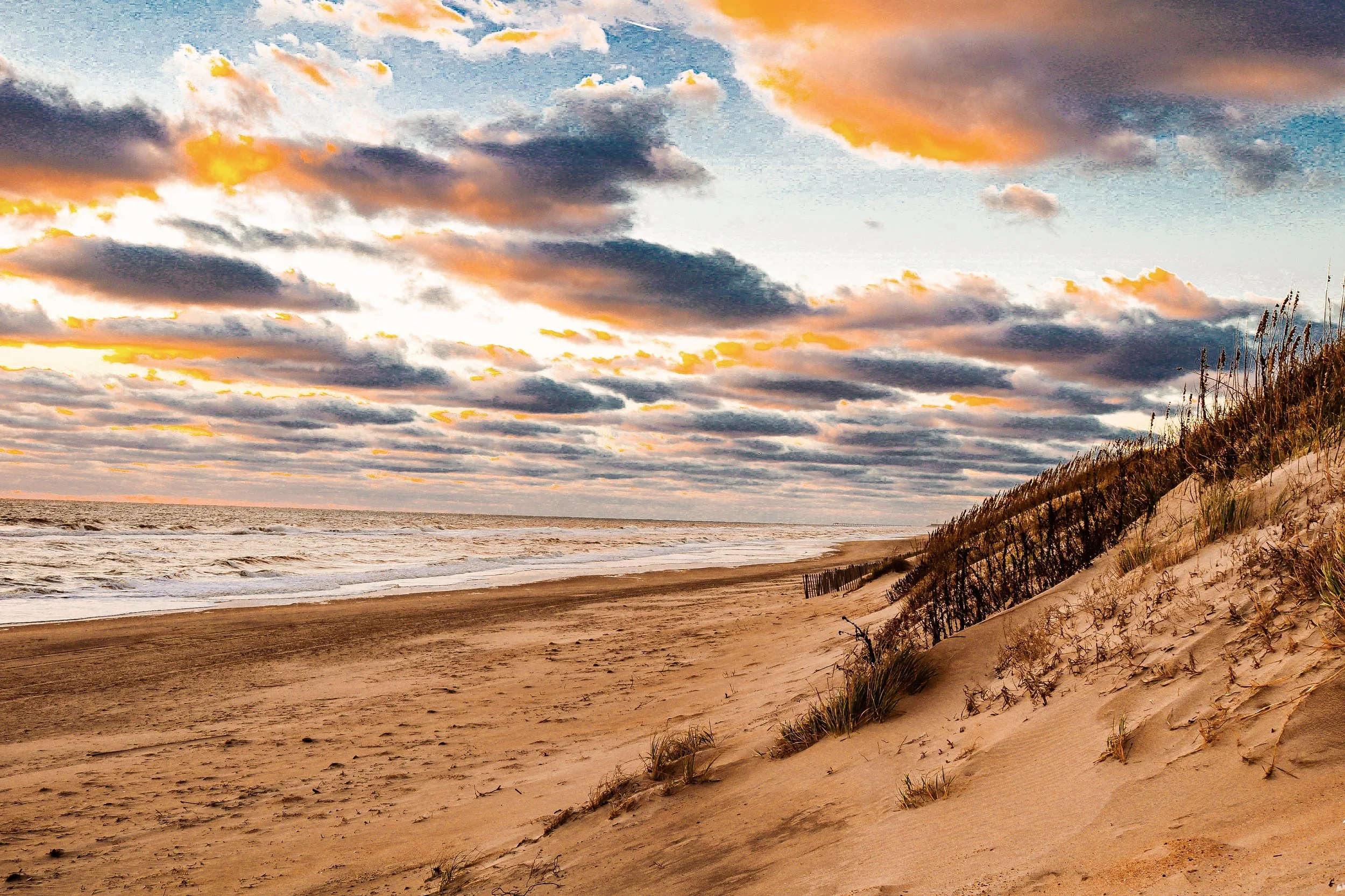 A sandy beach with dunes and a shoreline, cloudy sky with patches of orange and yellow sunset colors