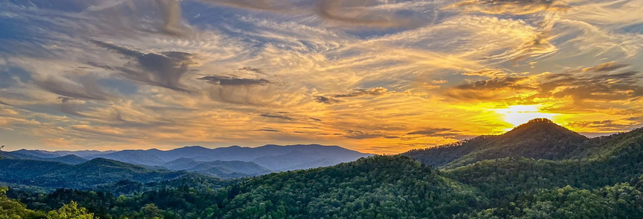 Sunset over a mountain range with colorful sky and layered hills covered in green trees.