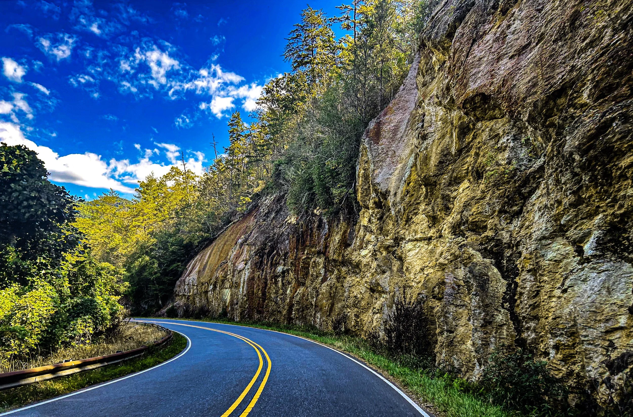 Curving mountain road next to rocky cliff with green trees and blue sky with clouds.