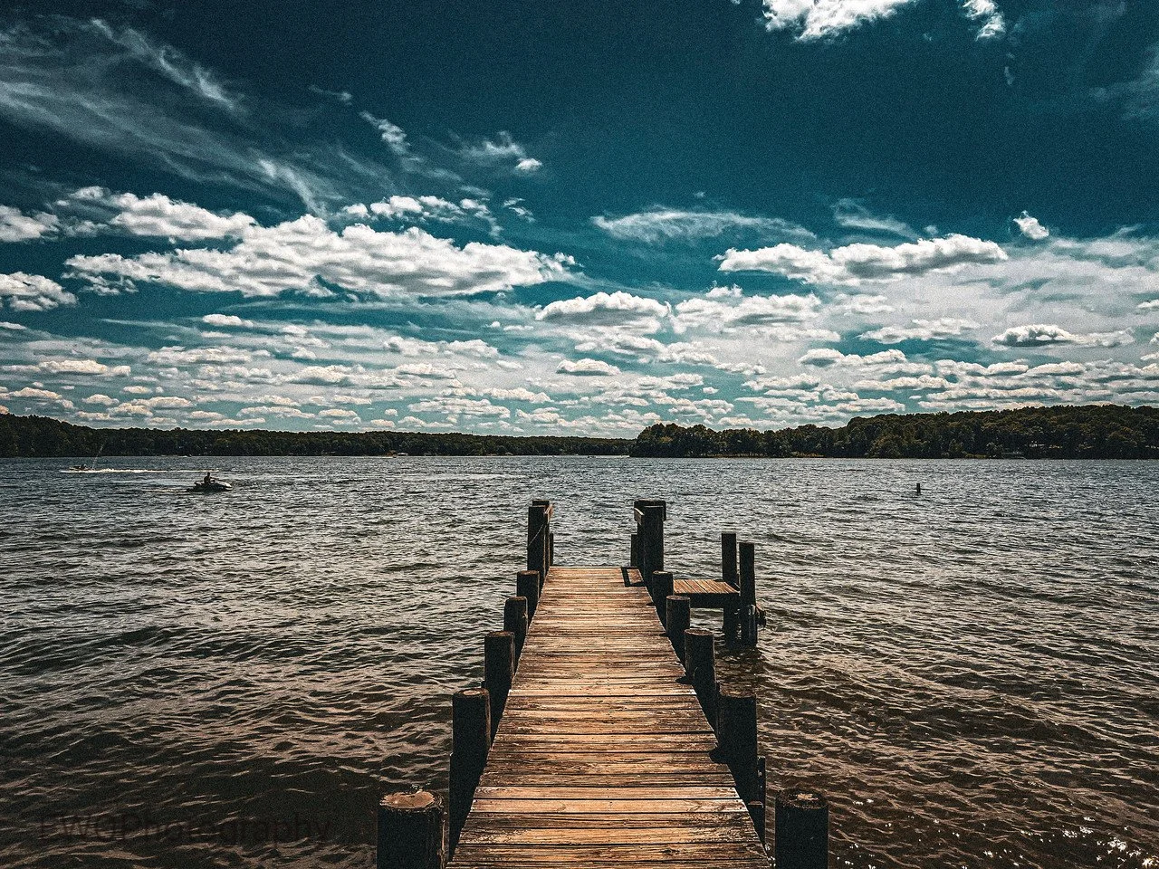 A wooden dock extending into a lake under a partly cloudy sky with some boats visible in the distance.