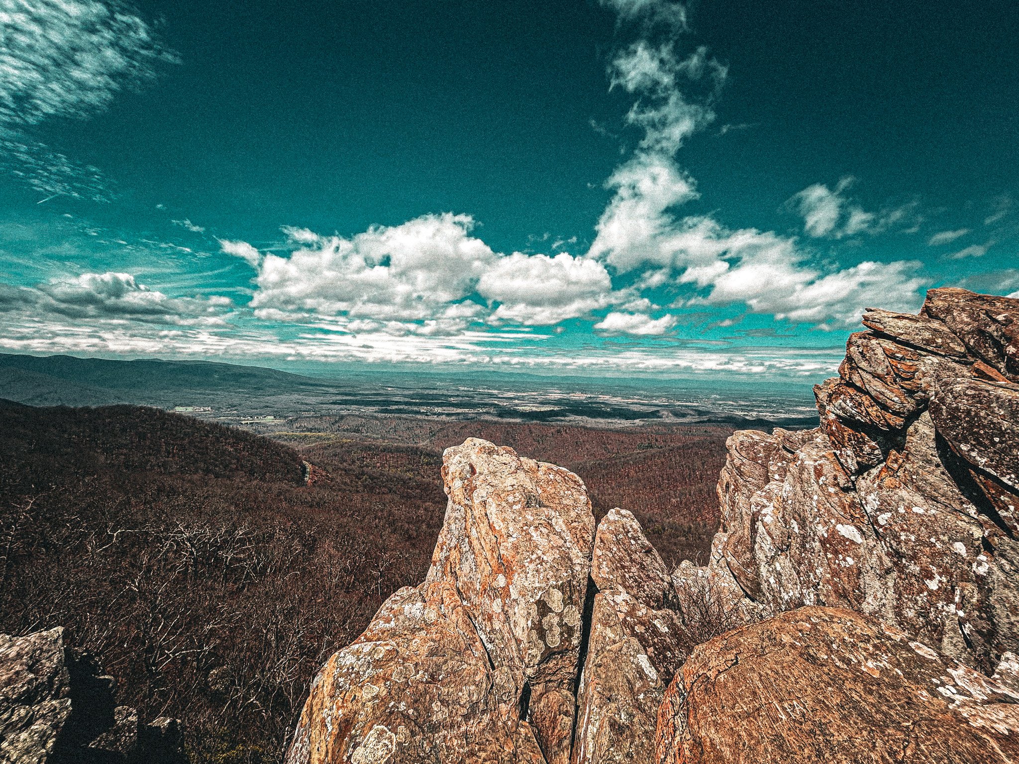 A panoramic view of a vast landscape with large, rugged rocks in the foreground, rolling hills covered with trees, and a sky filled with scattered clouds.