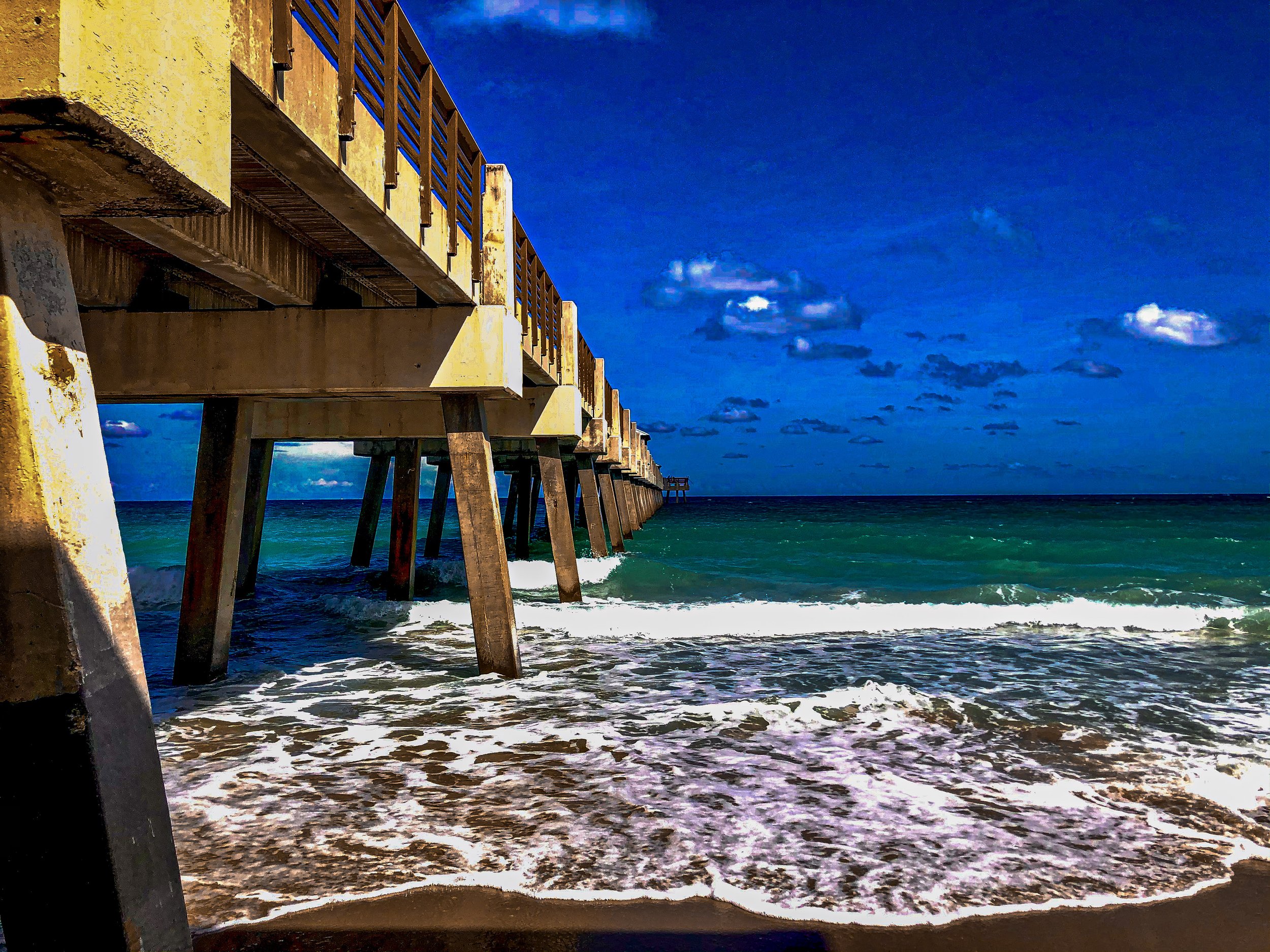View of a wooden pier extending over the ocean with waves crashing at the shore, under a blue sky with some clouds.