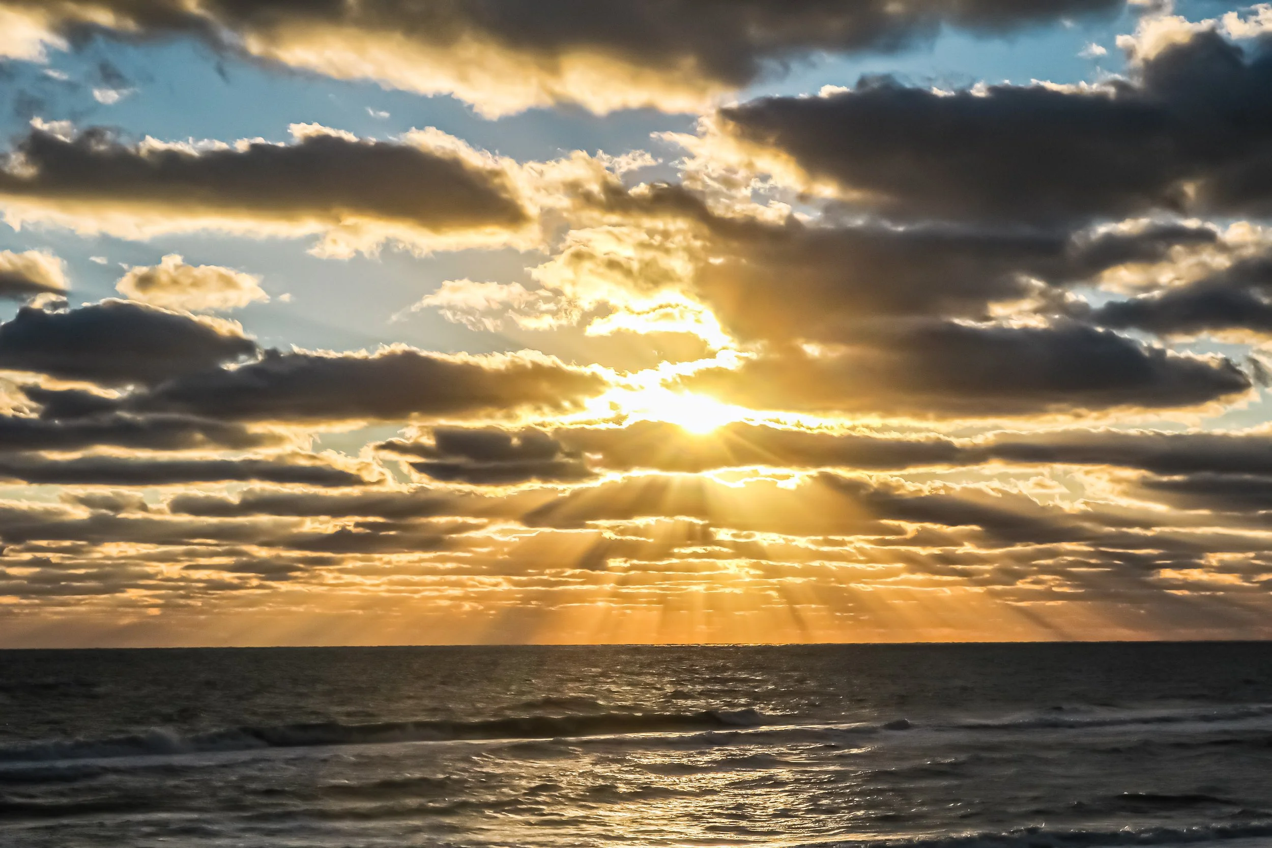 Sunset over the ocean with sun rays shining through dark clouds in the sky.