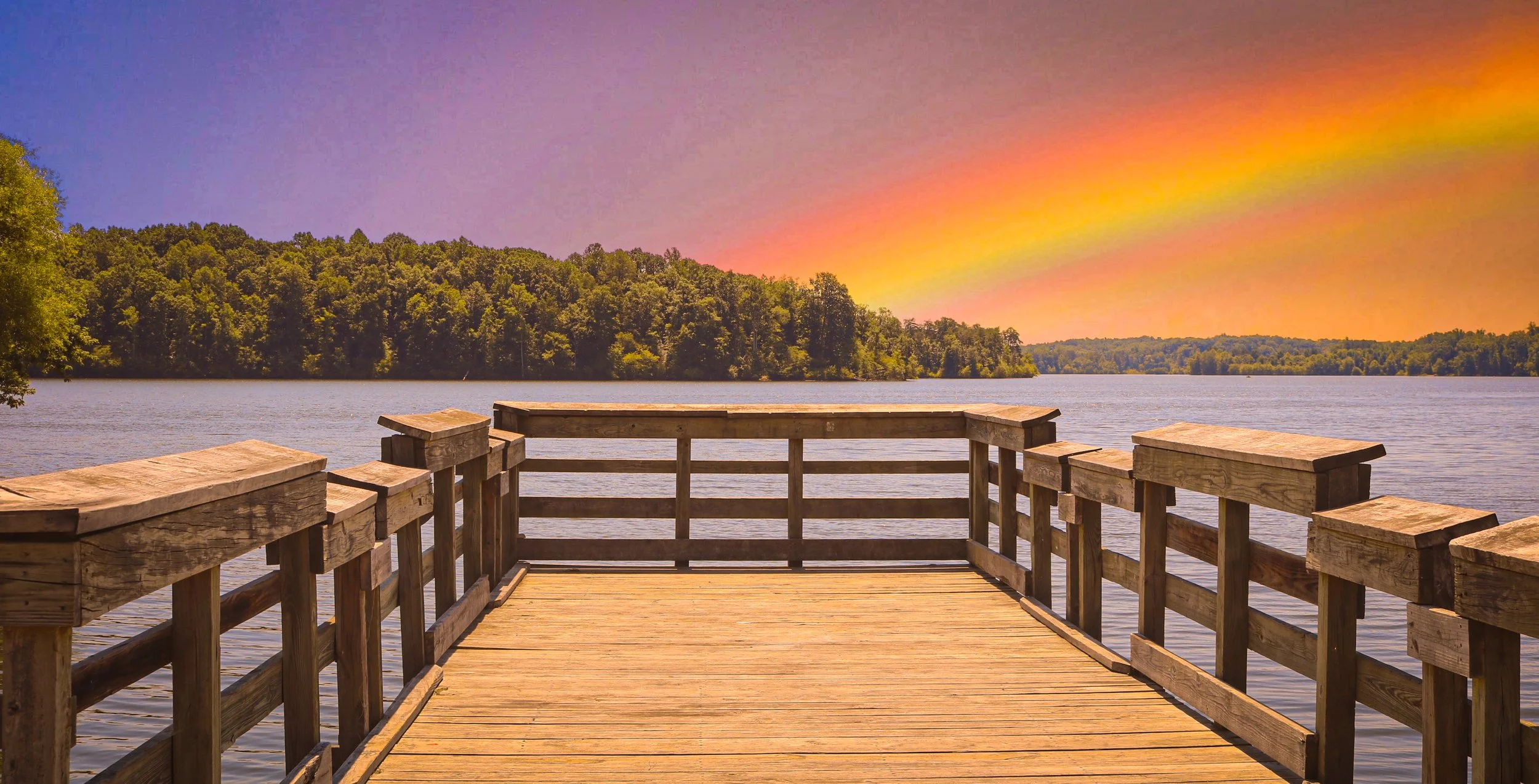 A wooden dock extends over a calm lake at sunset, with a colorful sky featuring shades of orange, yellow, pink, purple, and blue, and trees lining the distant shoreline.