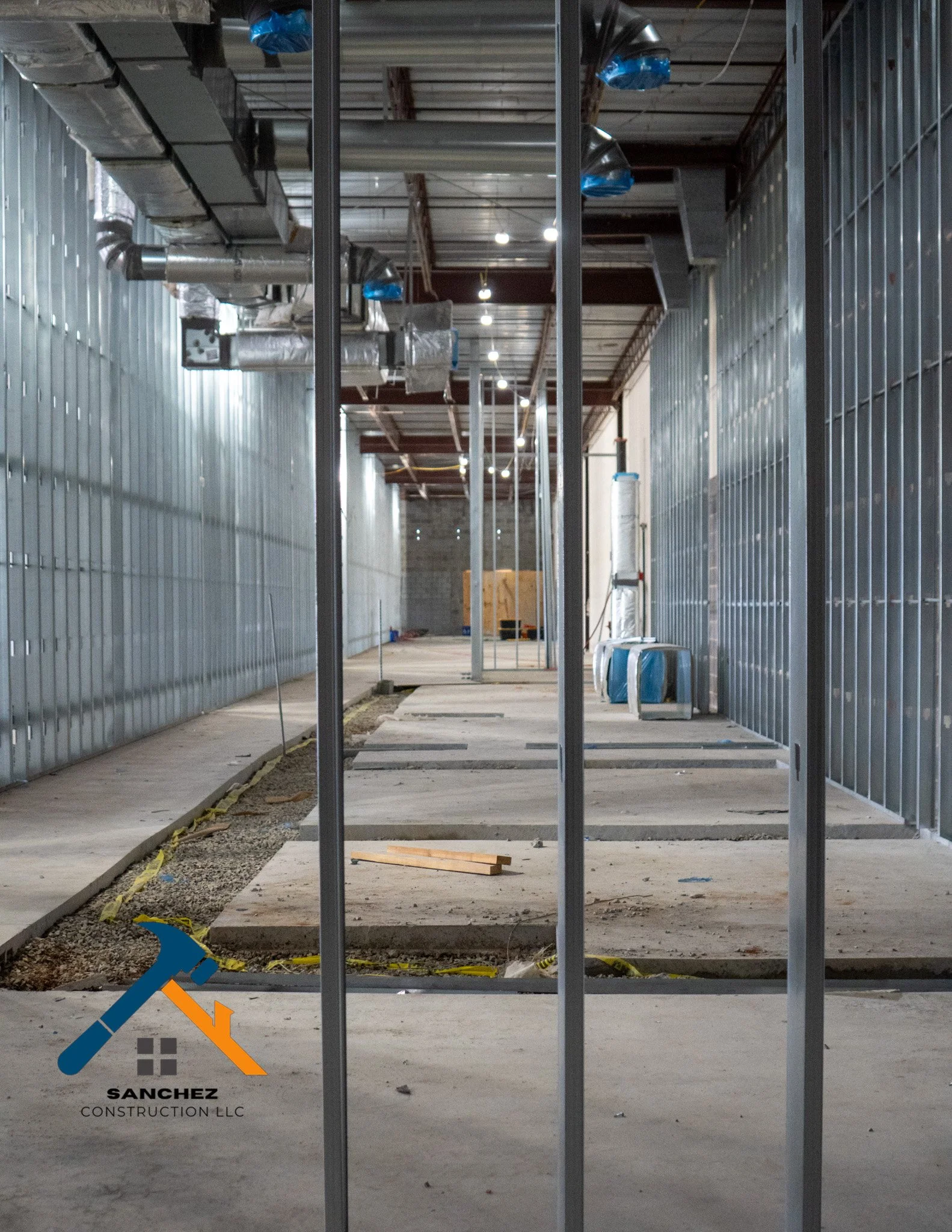 Interior view of a building under construction with steel framing, ductwork, and unfinished flooring. The Sanchez Construction LLC logo is in the bottom left corner.