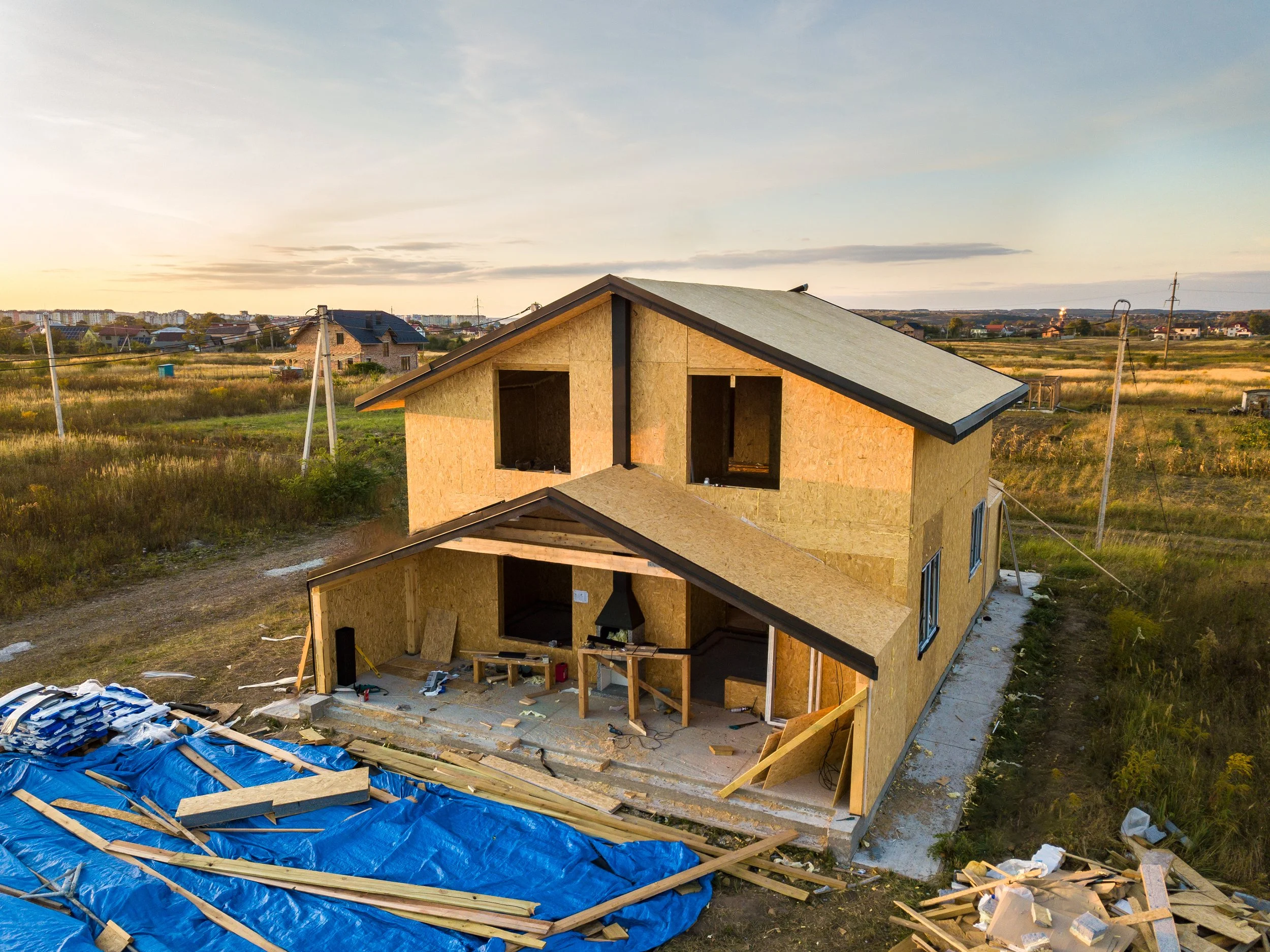 A house under construction with wooden walls and a sloped roof, surrounded by a grassy field, and several utility poles in the background at sunset.