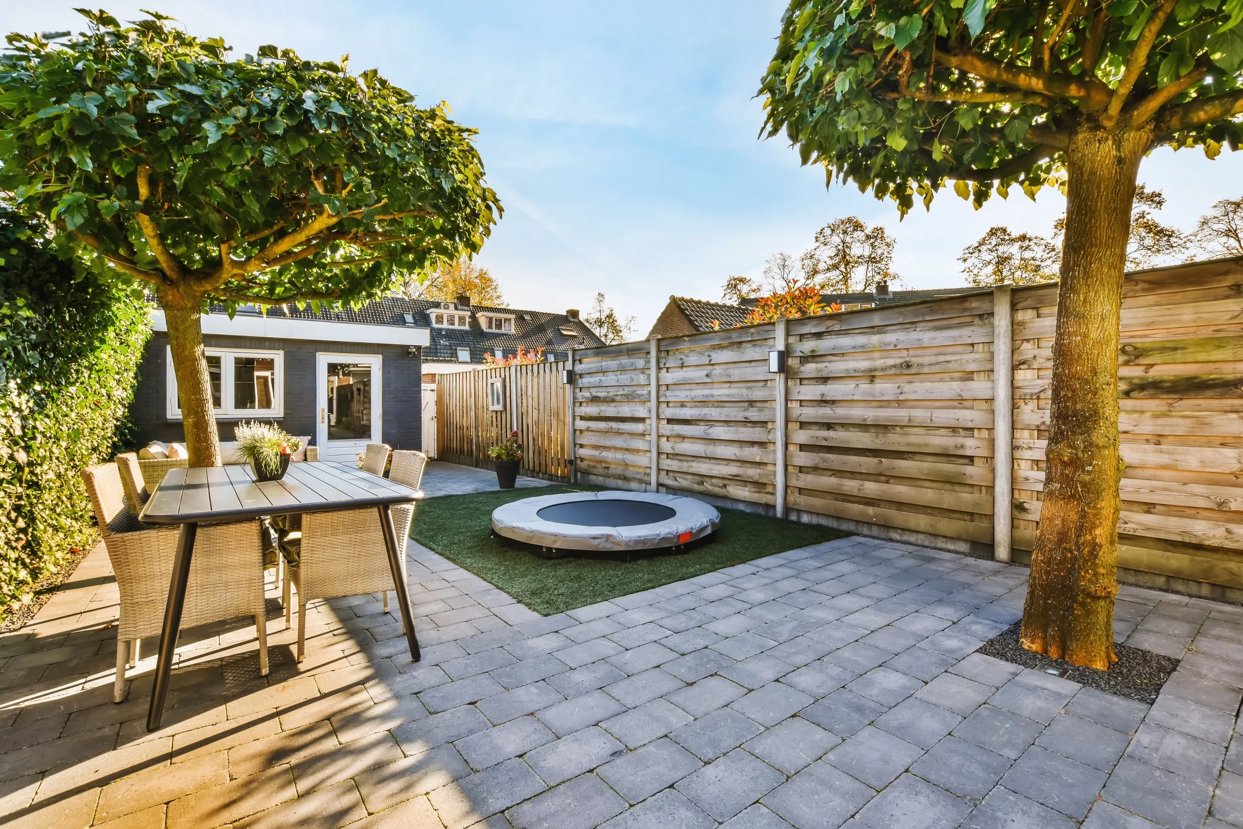 Backyard patio with two trees, a wooden fence, a potted plant, a round trampoline, a garden shed, and an outdoor dining table with chairs under tree shade