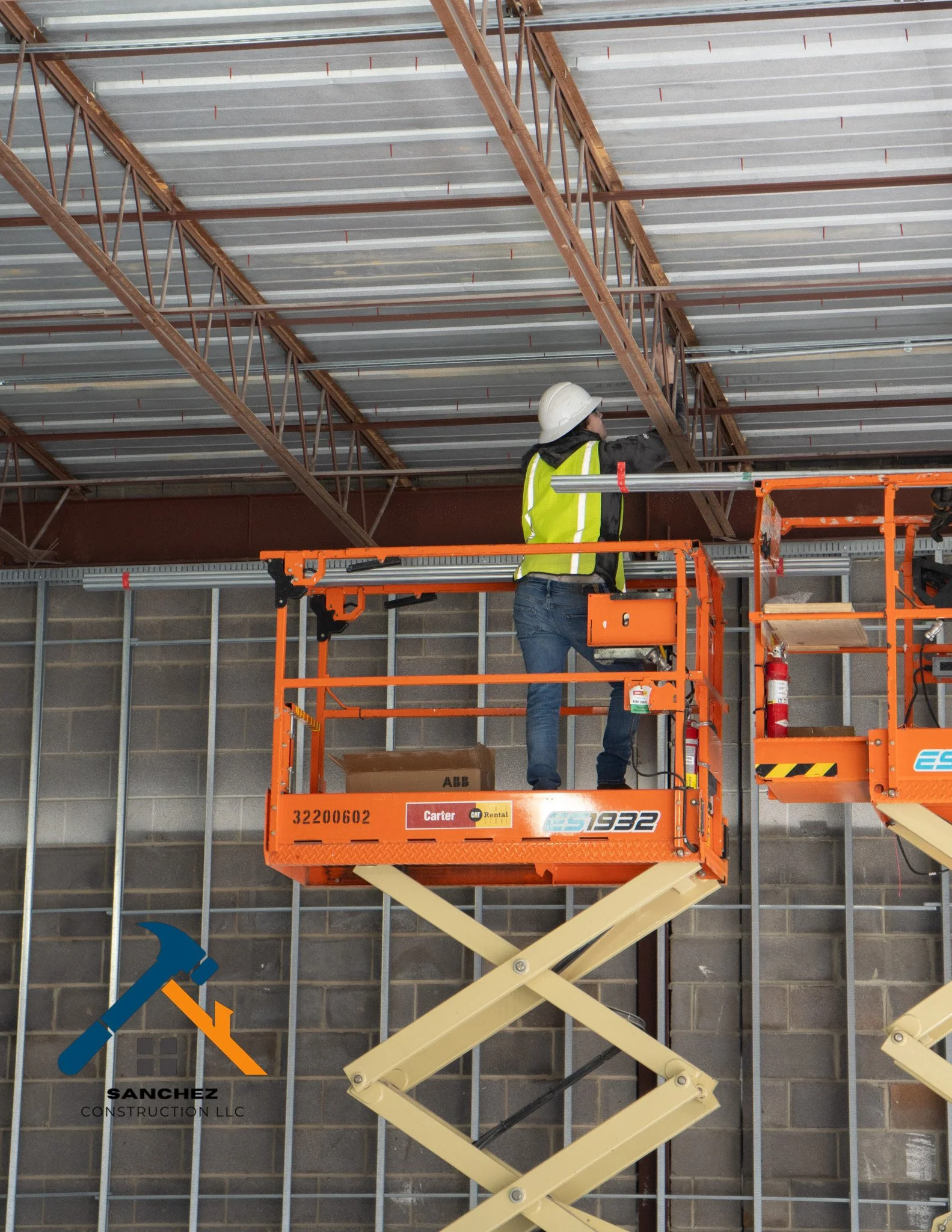 A construction worker in a yellow safety vest and white helmet is working on an indoor ceiling while standing on an orange scissor lift. The background shows gray brick walls and metallic framing for the interior structure.