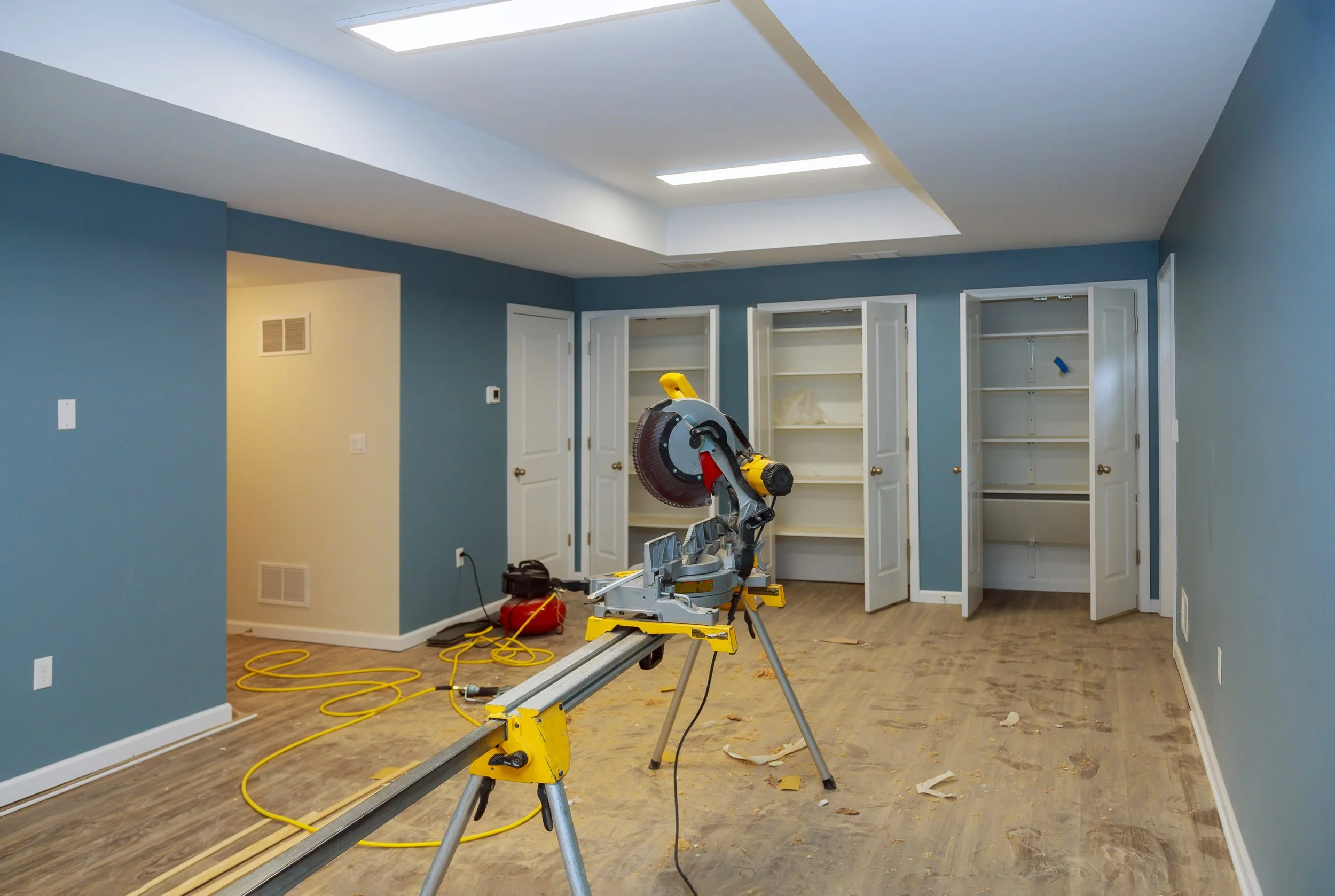 Room under renovation with blue walls, open white closets, hardwood floor, and a miter saw on a stand in the center, with yellow extension cords and construction tools visible.