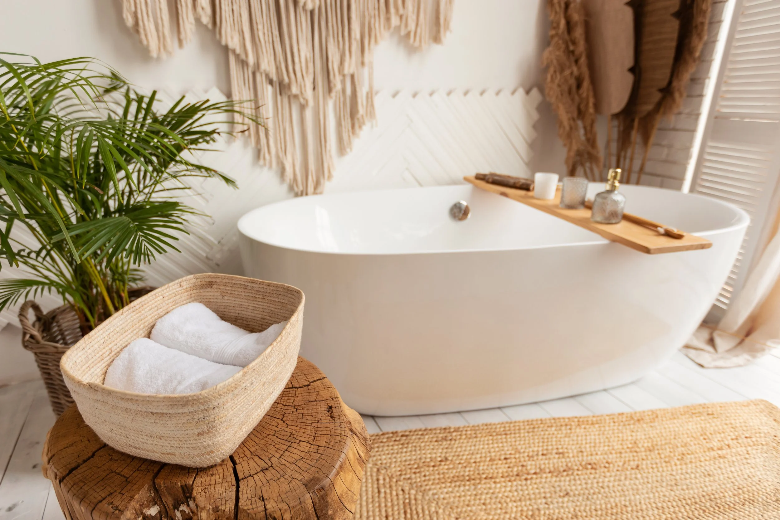Bathroom with a standalone white bathtub, a wooden tray with candles and skincare bottles, a wicker basket with towels, a potted plant, and woven decor on the wall.