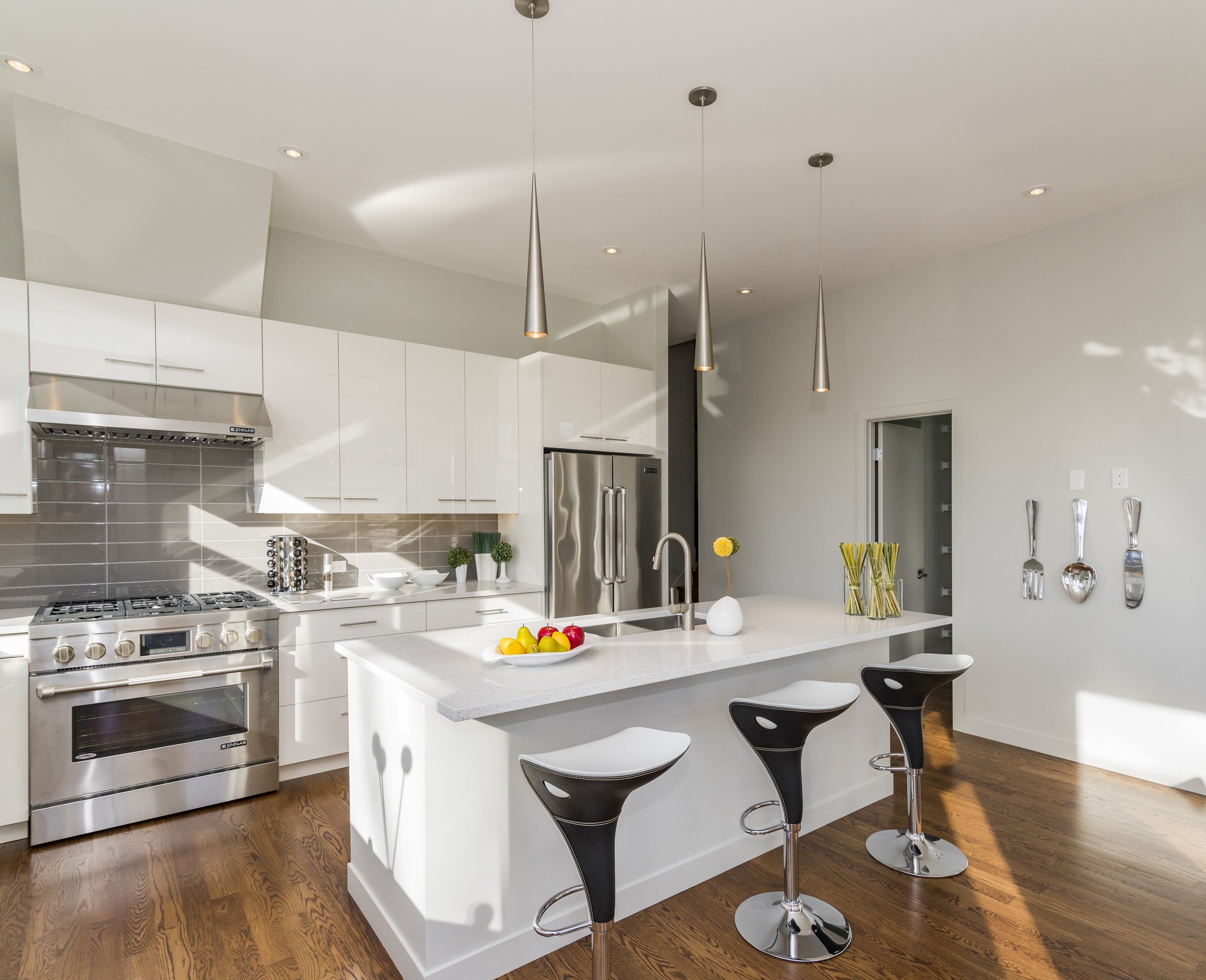 Modern white kitchen with stainless steel appliances, a kitchen island with bar stools, and decorative utensils on the wall.