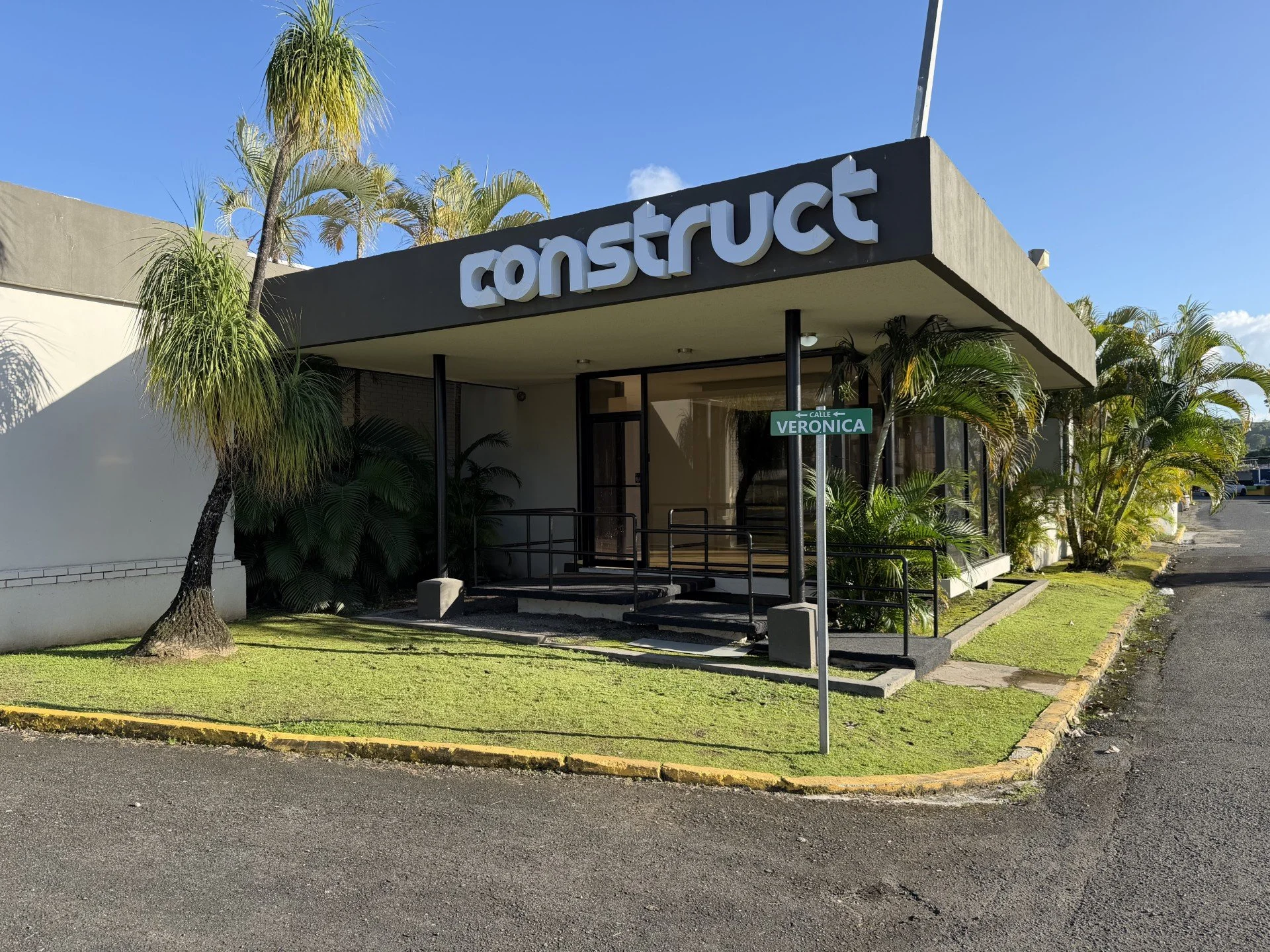 Exterior of a building with a large sign that says 'construct.' The building is surrounded by palm trees and has a sidewalk leading to the entrance with a ramp. There's a street sign that reads 'Calle Veronica'.