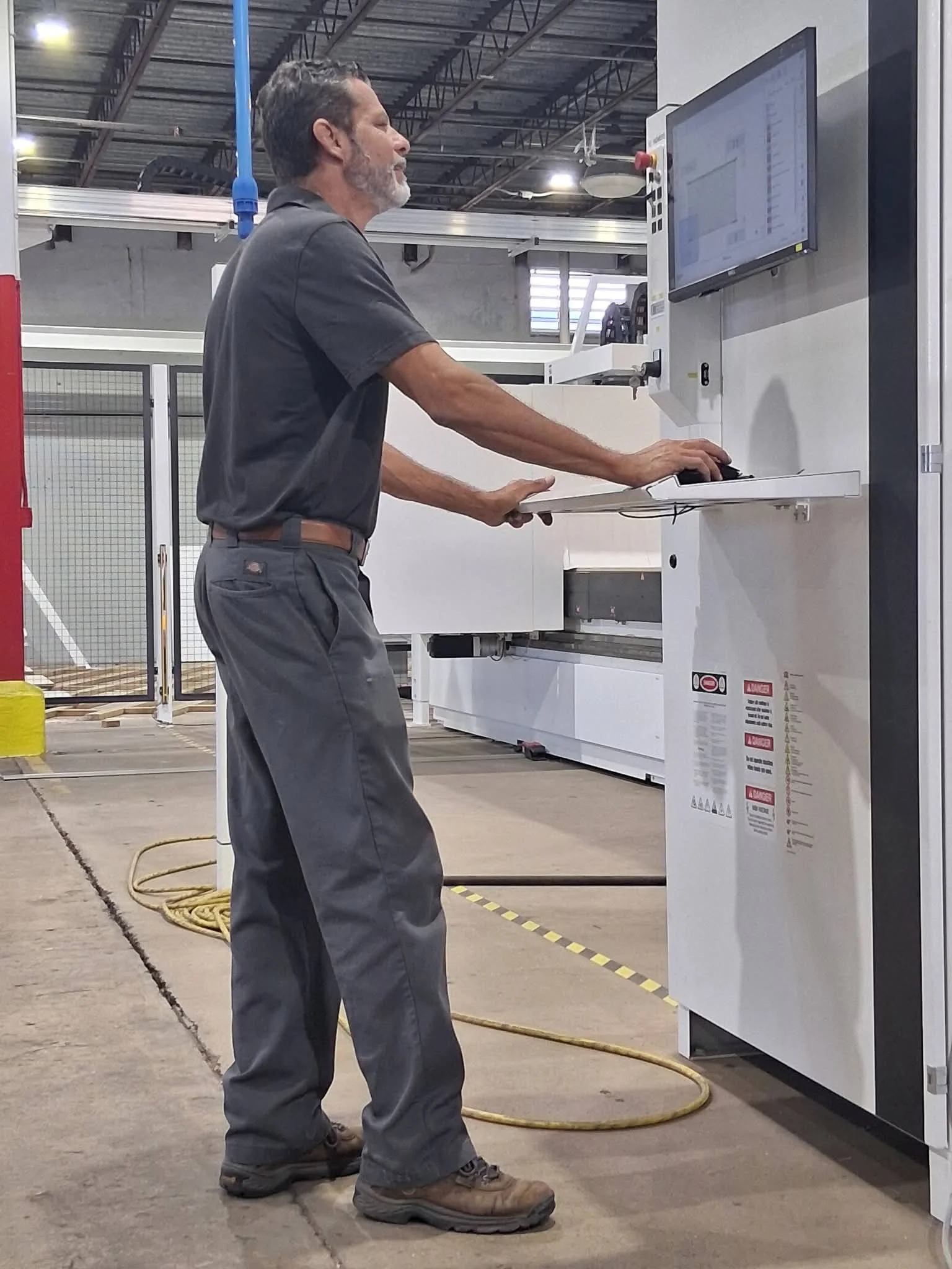 A man operating an industrial machine and computer in a large warehouse. He is standing and using the computer interface on the machine.