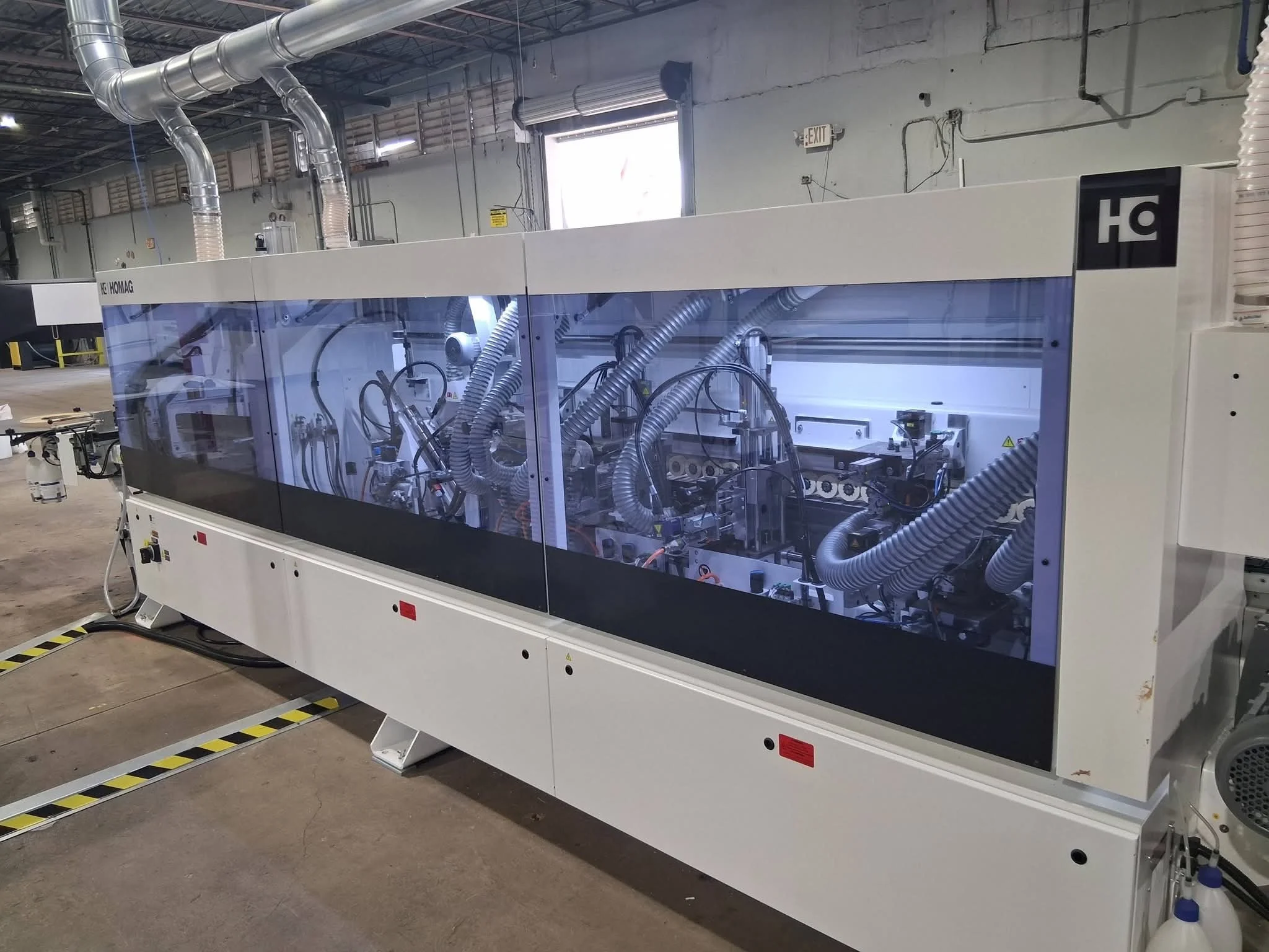 Large industrial machine with transparent panels, multiple hoses, and mechanical components inside, located in a warehouse or factory.