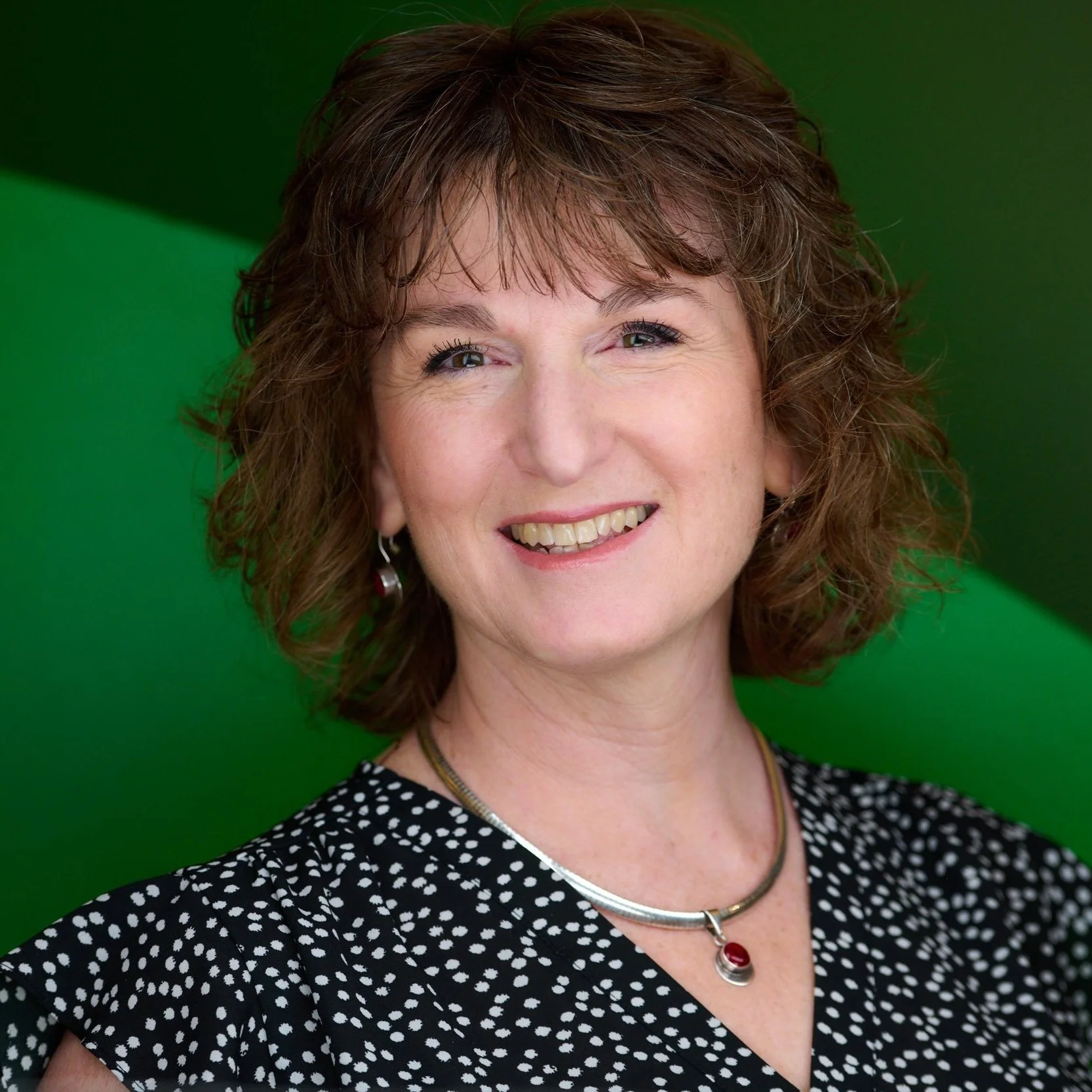 A woman with shoulder-length curly brown hair smiling, wearing a black and white patterned top, a silver necklace with a red pendant, and silver earrings, standing against a green background.