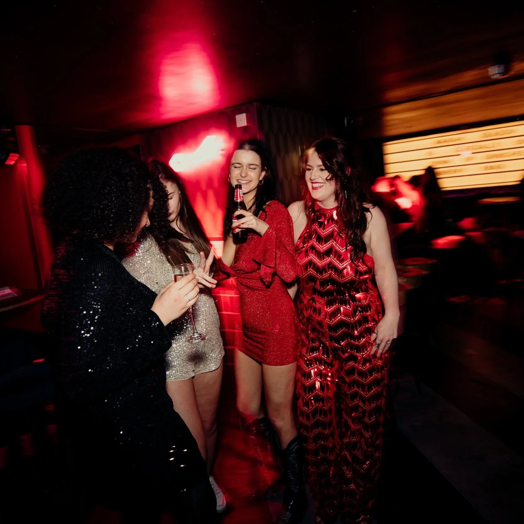 Group of women laughing and talking at a party, holding drinks, dressed in red and black outfits, with a blurred bar or lounge background.