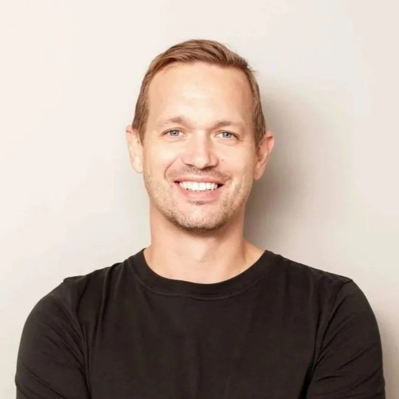 Portrait of a smiling man with crossed arms, wearing a black t-shirt, standing against a plain light-colored background.