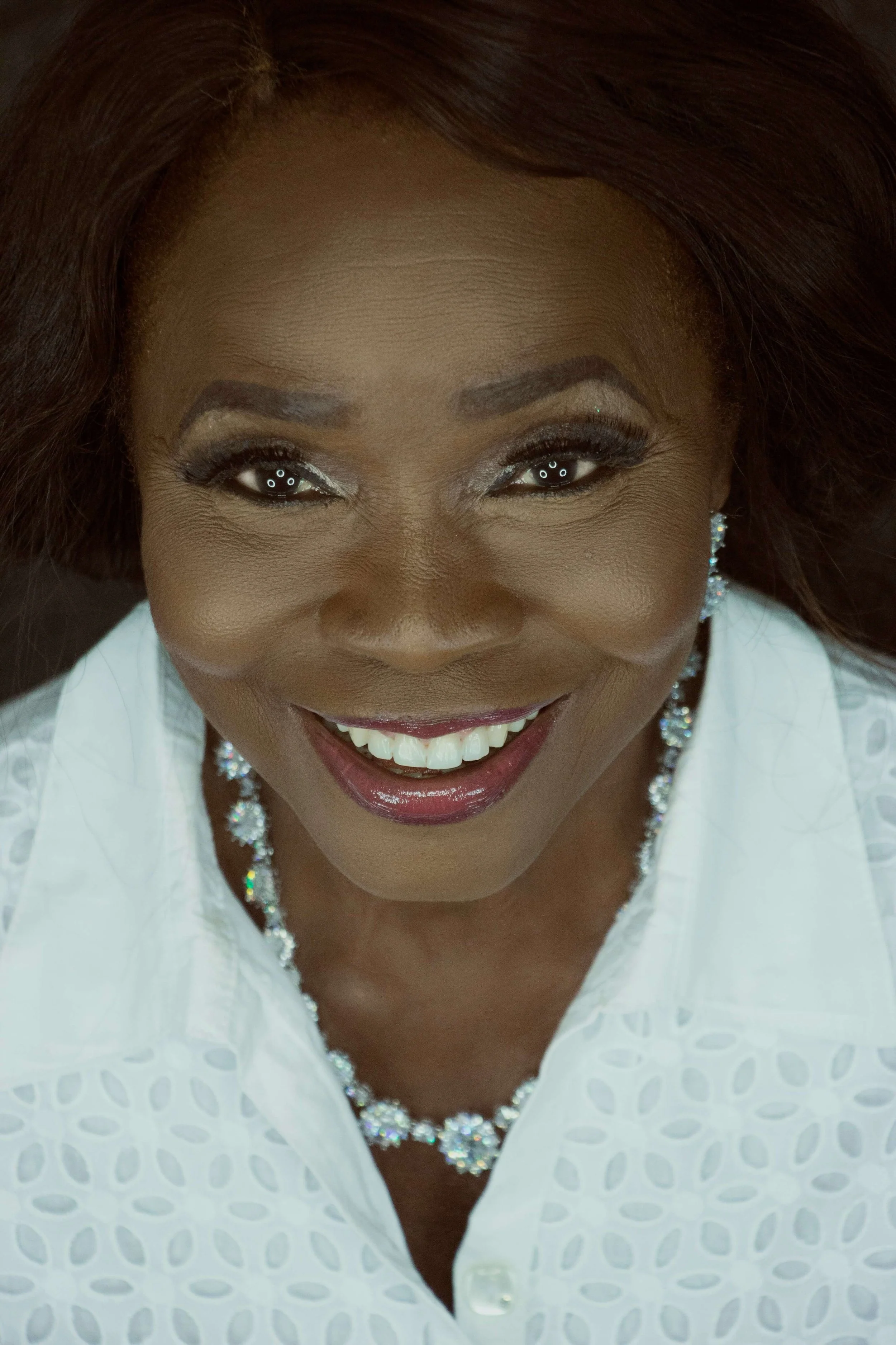 Close-up of a smiling  woman with brown hair, wearing a white blouse and sparkling jewelry, looking directly at the camera.