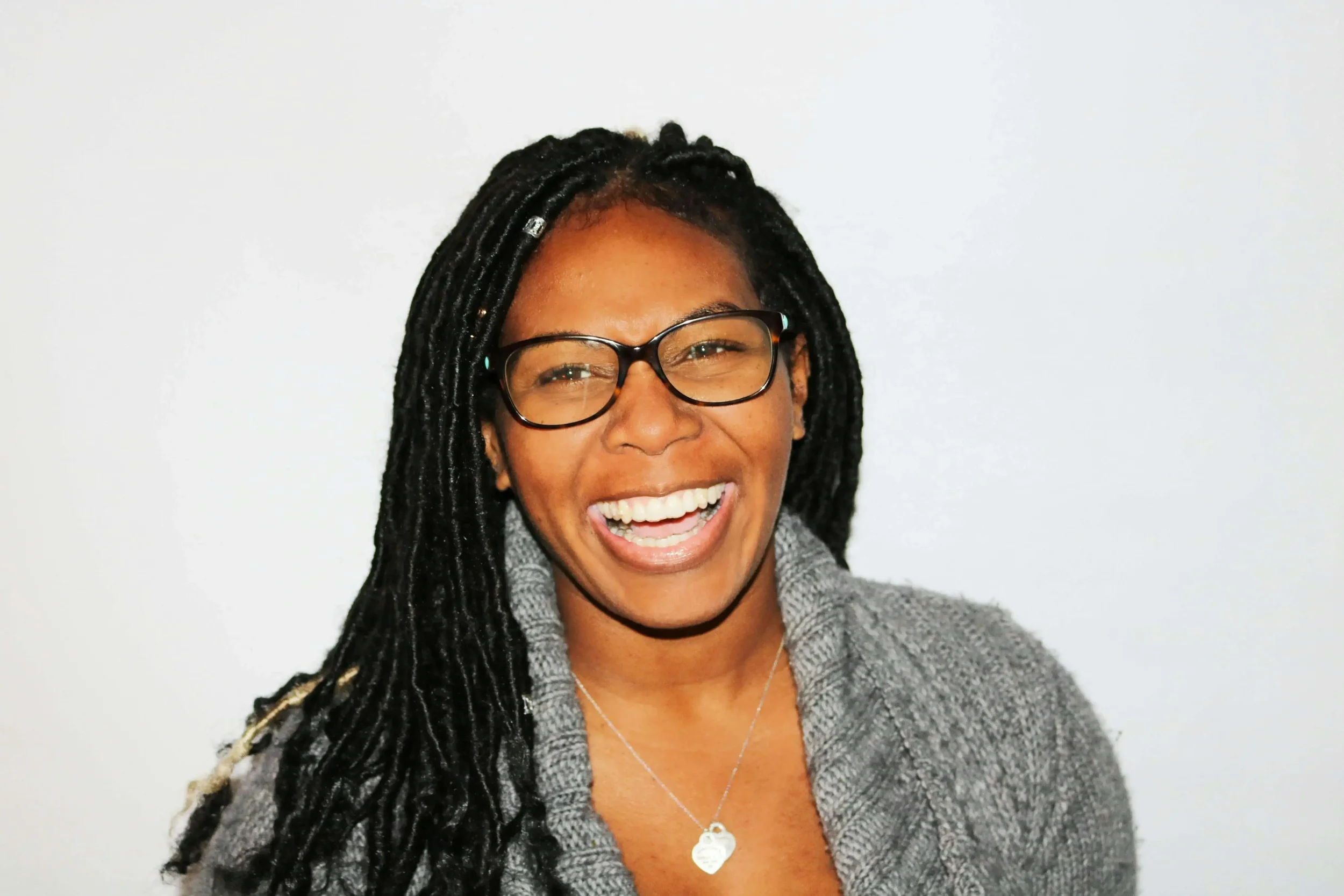 A smiling woman with glasses and dreadlocks wearing a gray sweater and a heart-shaped necklace against a plain white background.