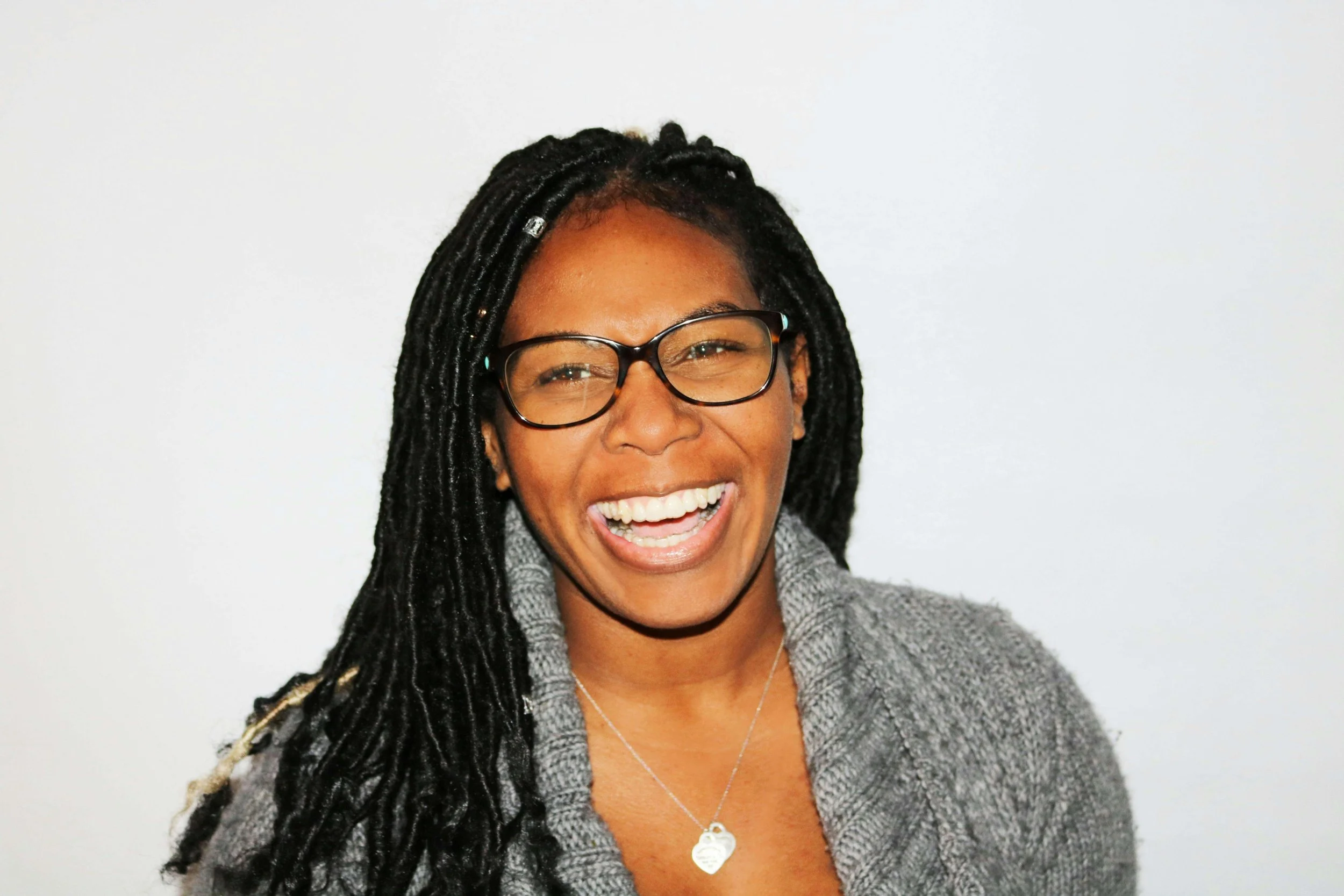 A woman with glasses and long black dreadlocks smiling against a plain white background.