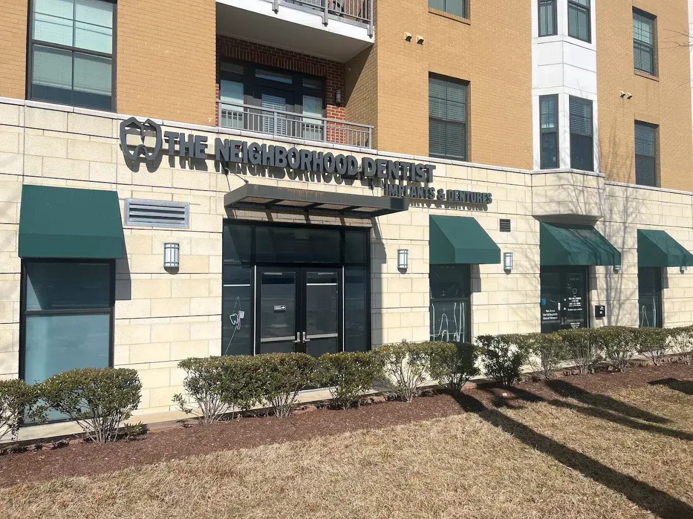 Exterior of a dental office called The Neighborhood Dentist with green awnings, windows, and bushes in front.