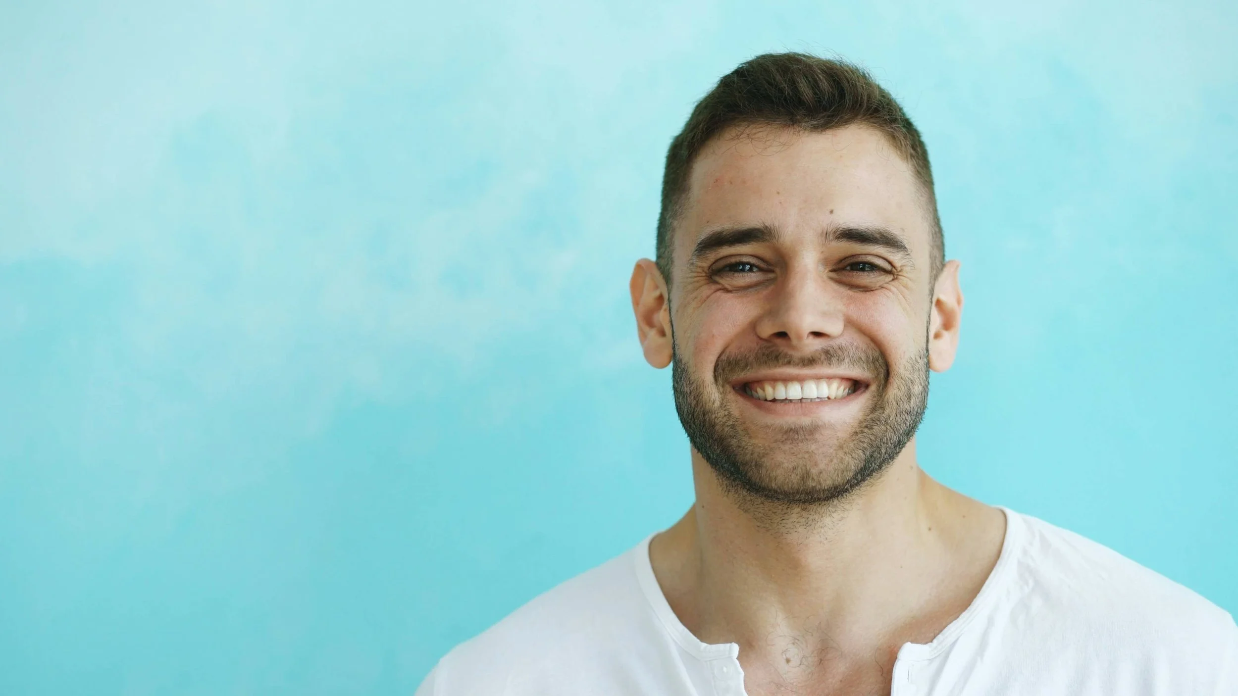 Smiling young man with short brown hair and a beard, wearing a white shirt, against a light blue background.