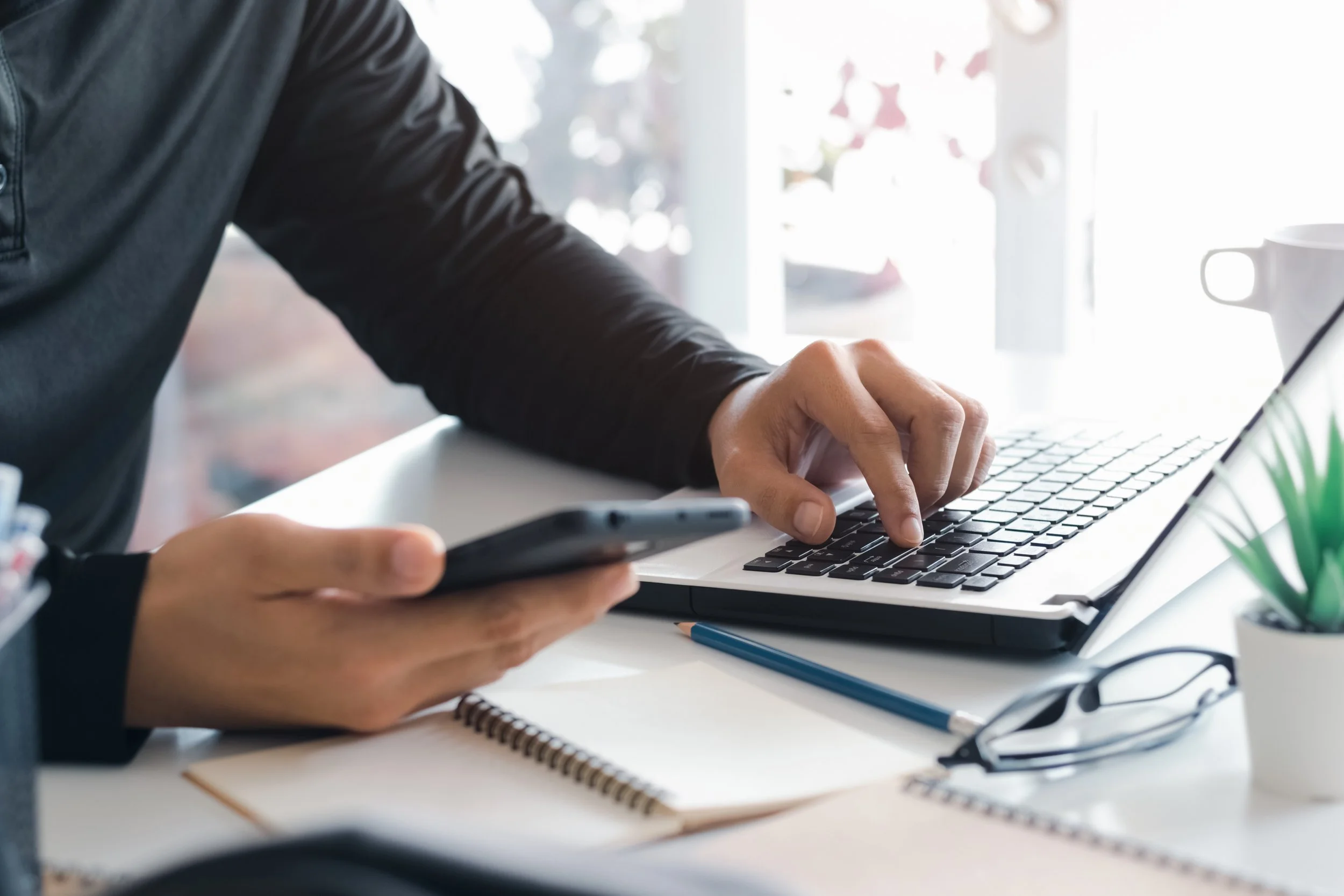 Person working on laptop and holding smartphone at a desk with notebook, glasses, pen, and potted plant.