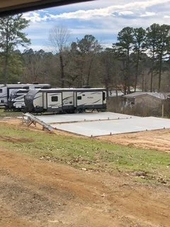 Several RV trailers parked on a concrete pad in a rural area with trees and cloudy sky in the background.
