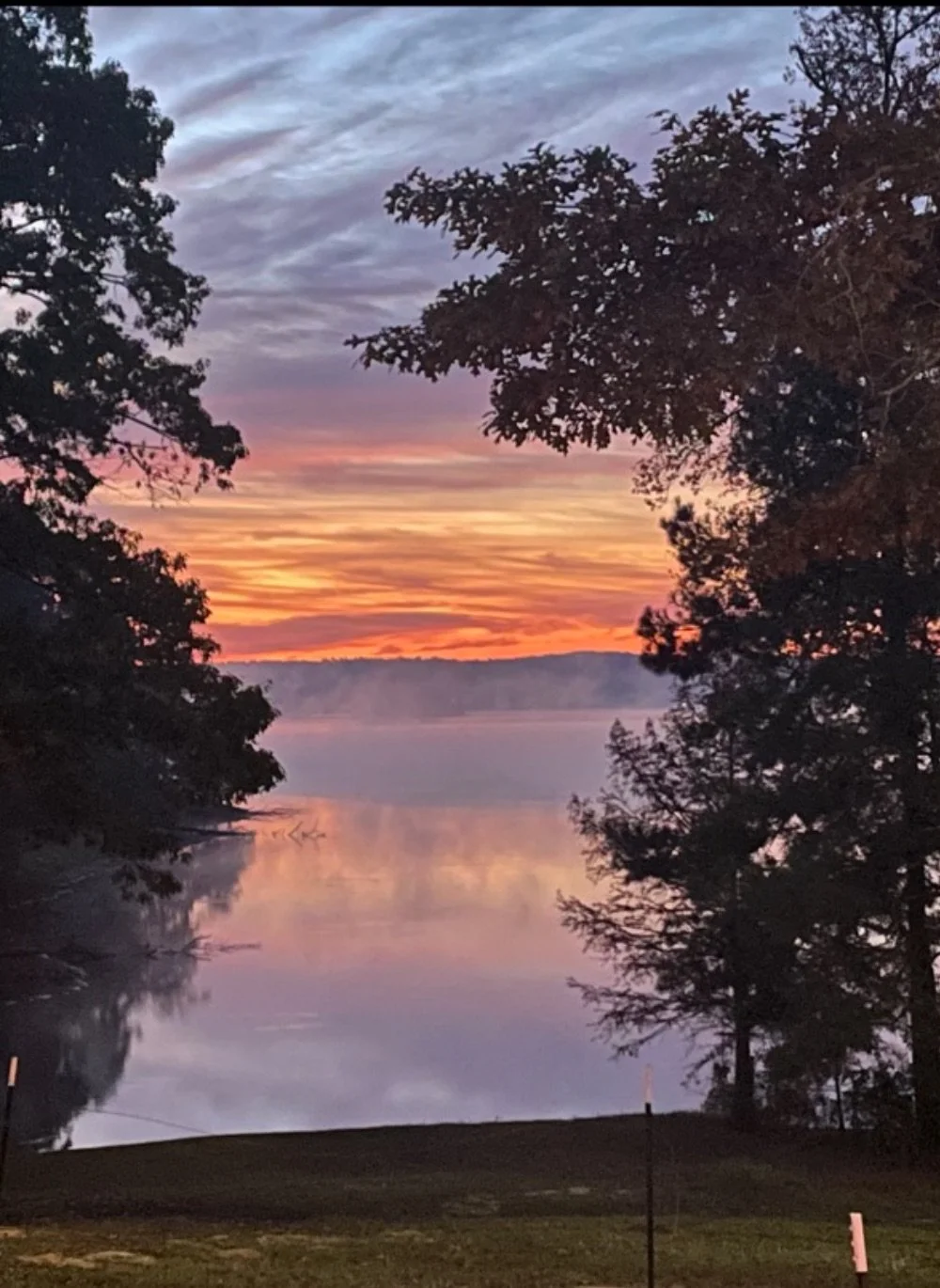 A serene lake during sunset with colorful sky and orange, pink, purple clouds, framed by trees on both sides.