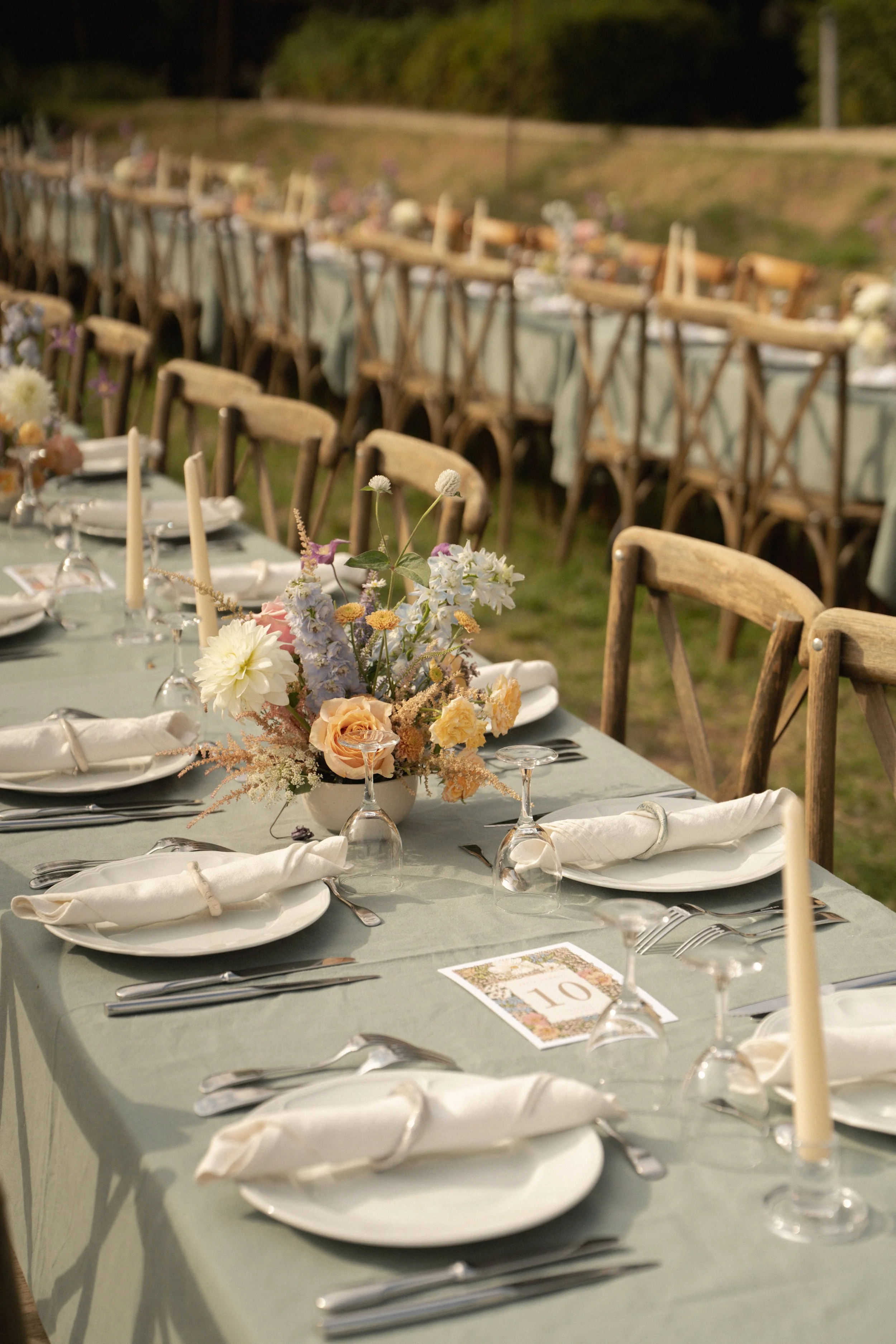 Outdoor dining table set for a wedding, featuring floral centerpieces, candles, white plates, napkins, and glassware, with wooden chairs and a long table extending outside.