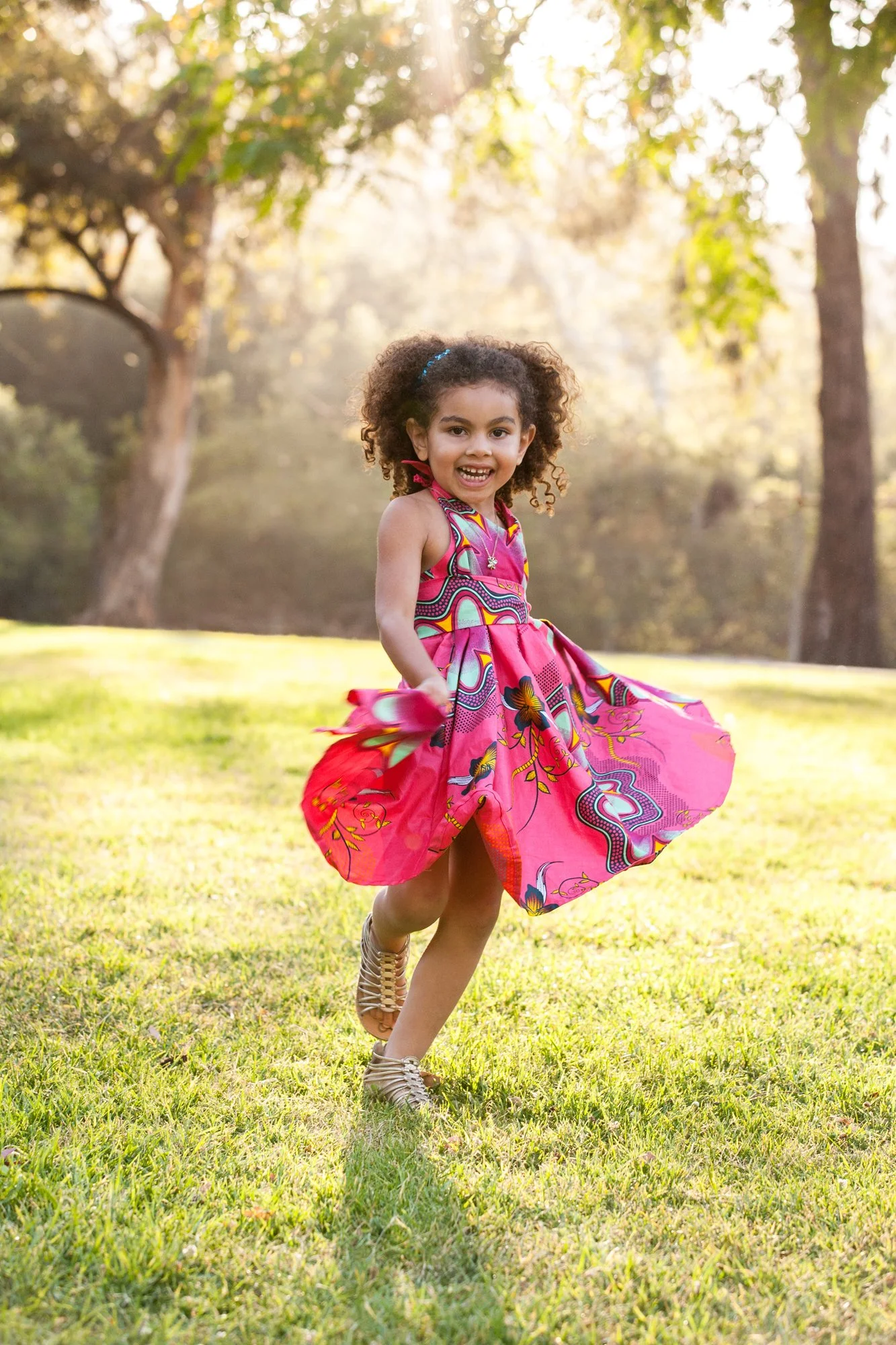 A young girl running on a grassy field, smiling, wearing a colorful pink dress and gold shoes, with trees and sunlight in the background.
