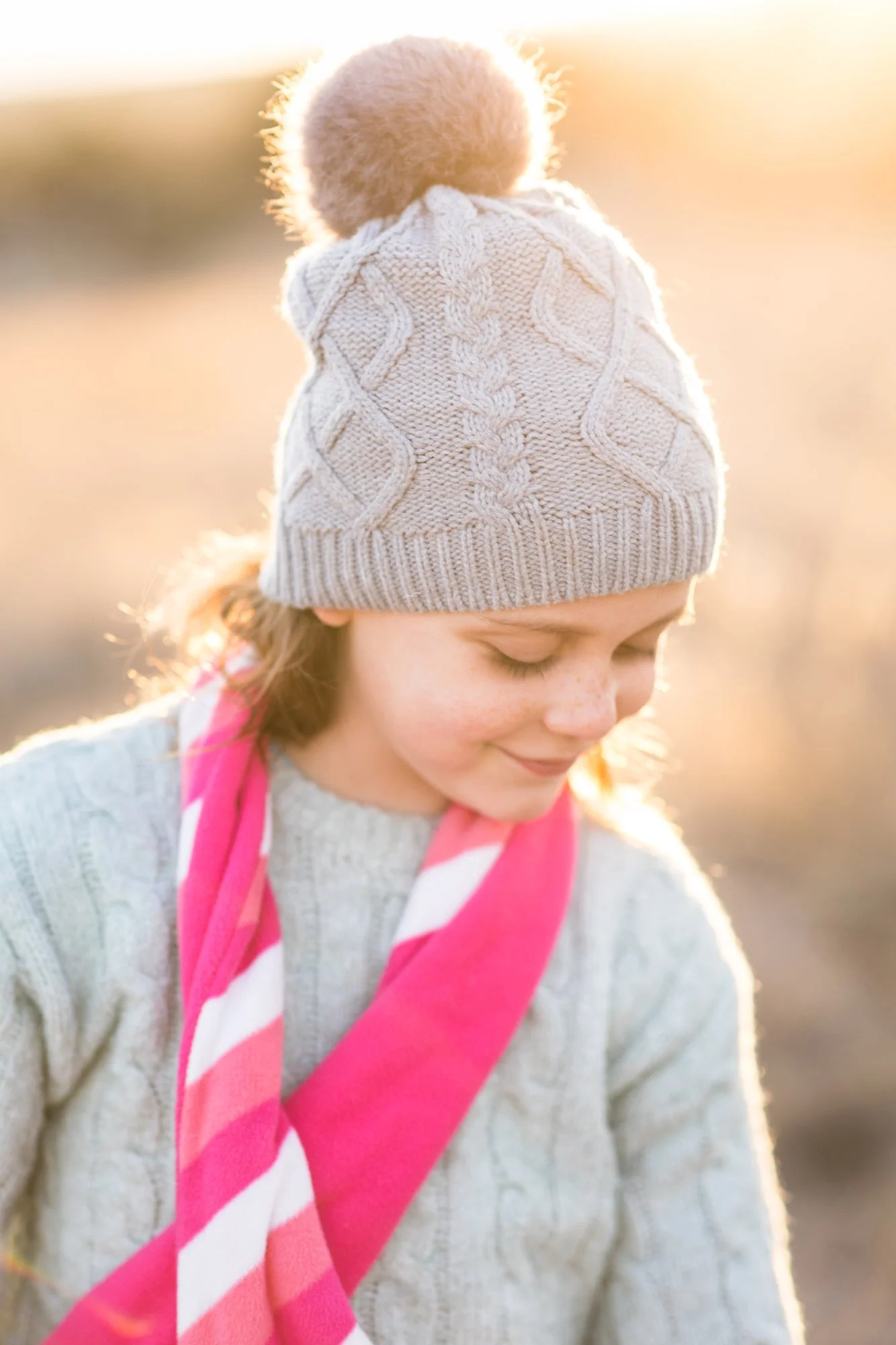 A young girl with freckles wears a knit hat with a pom-pom and a pink-and-white scarf, standing outdoors with sunlight in the background.