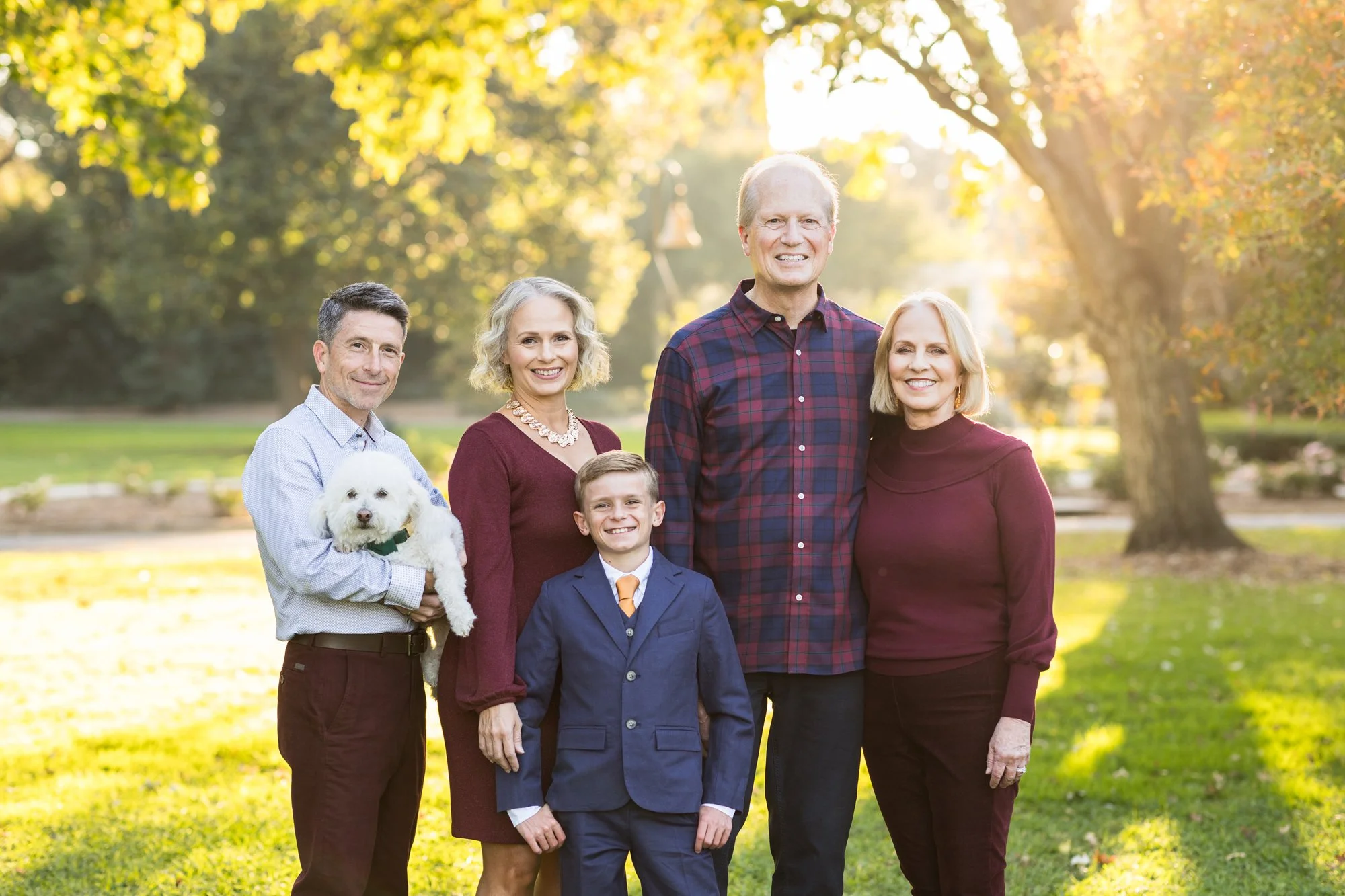 A family of five posing outdoors in a park during fall, including a man, woman, young boy, two middle-aged adults, and a dog.