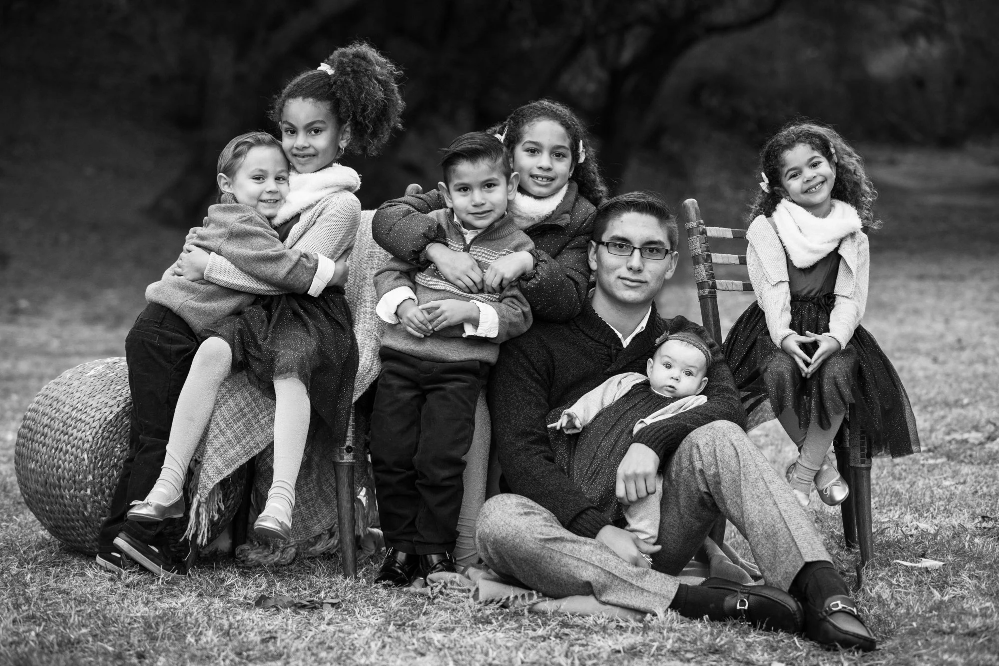 A diverse group of seven children and one adult posing outdoors for a black and white photo, some sitting, some leaning, with a tree and grass background.