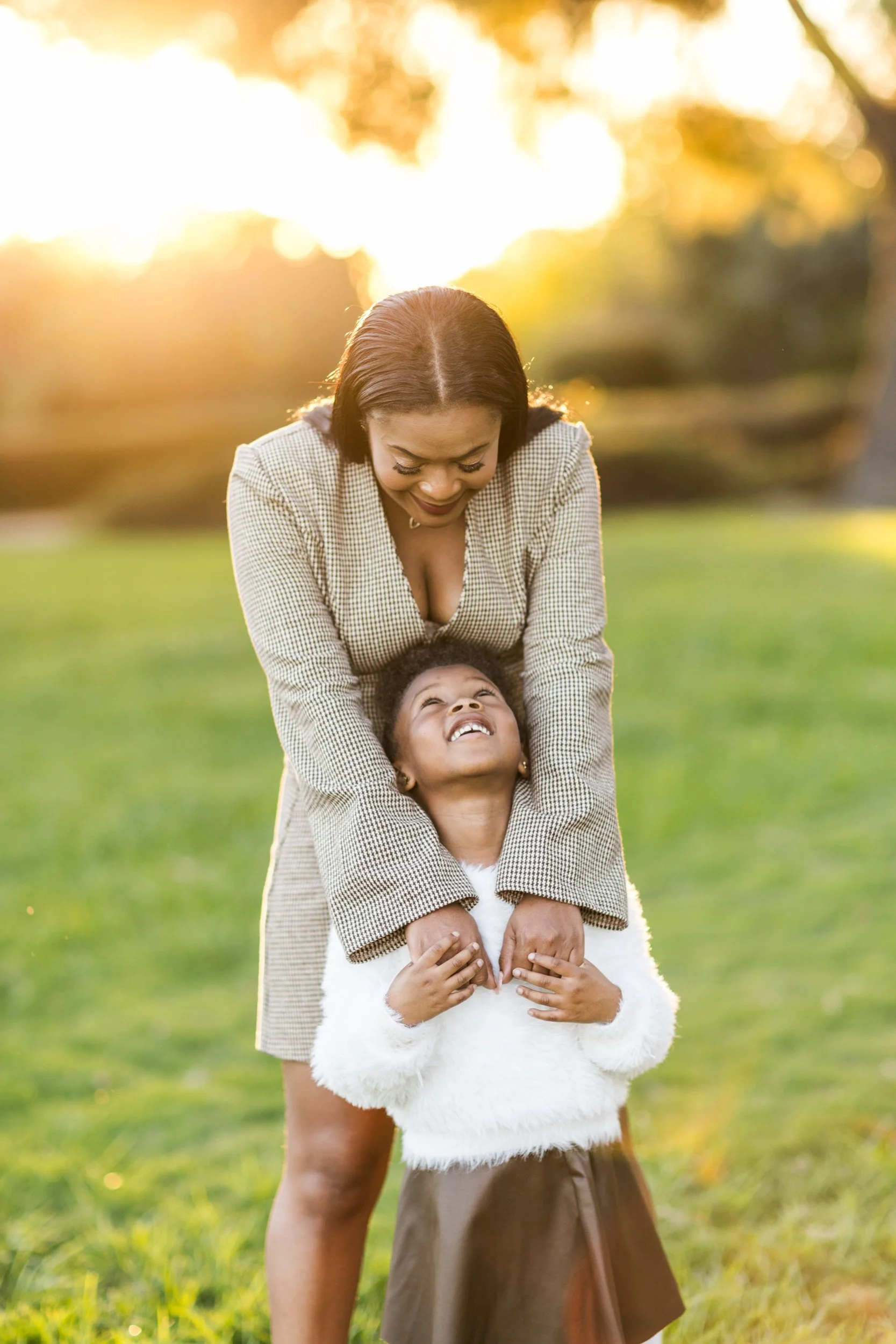 A woman and a young girl smiling and playing together outdoors in a park during sunset.