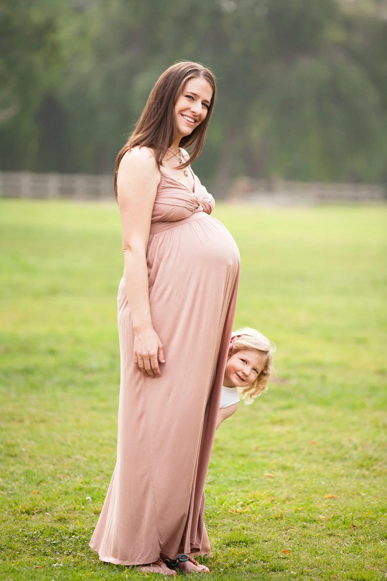 A pregnant woman in a pink dress smiling and standing on a grassy field with her young daughter peeking out from underneath her dress.