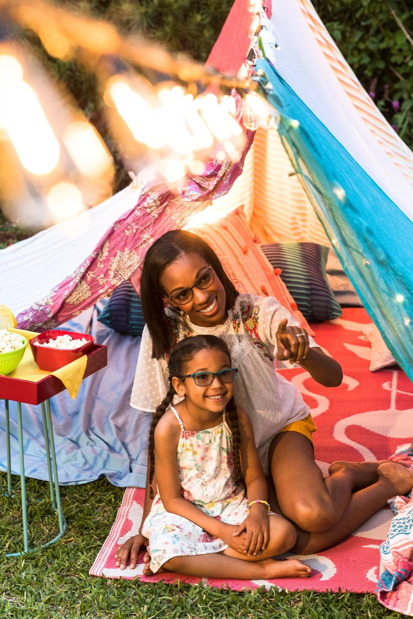 A woman and a girl sitting on a blanket inside a colorful fabric fort with string lights, smiling and enjoying a picnic outdoors.