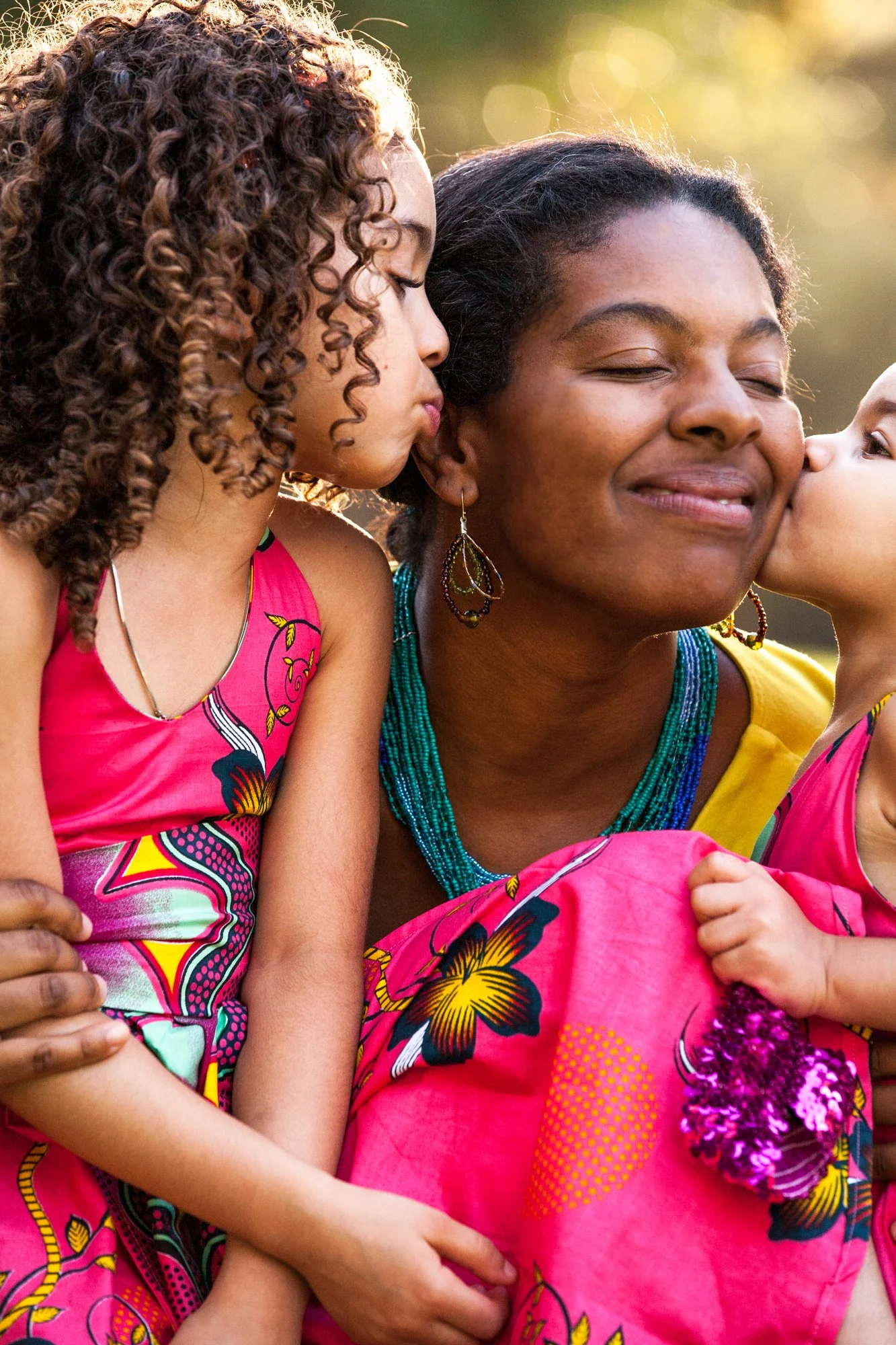 A woman with a joyful expression being kissed on the cheek by two young girls, outdoors with sunlight and greenery in the background.
