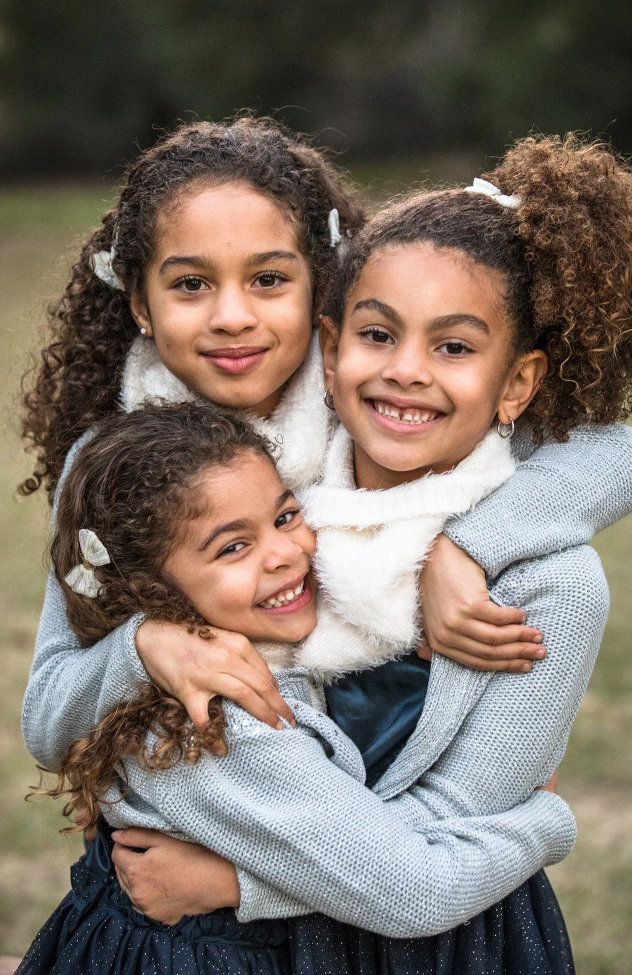 Three young girls with curly hair smiling and hugging outdoors.