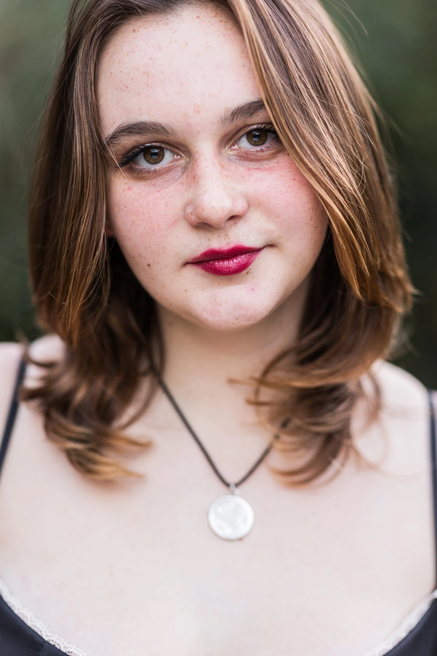 Close-up of a young woman with shoulder-length brown hair, wearing makeup with red lipstick, a black top, and a circular pendant necklace, outdoors.